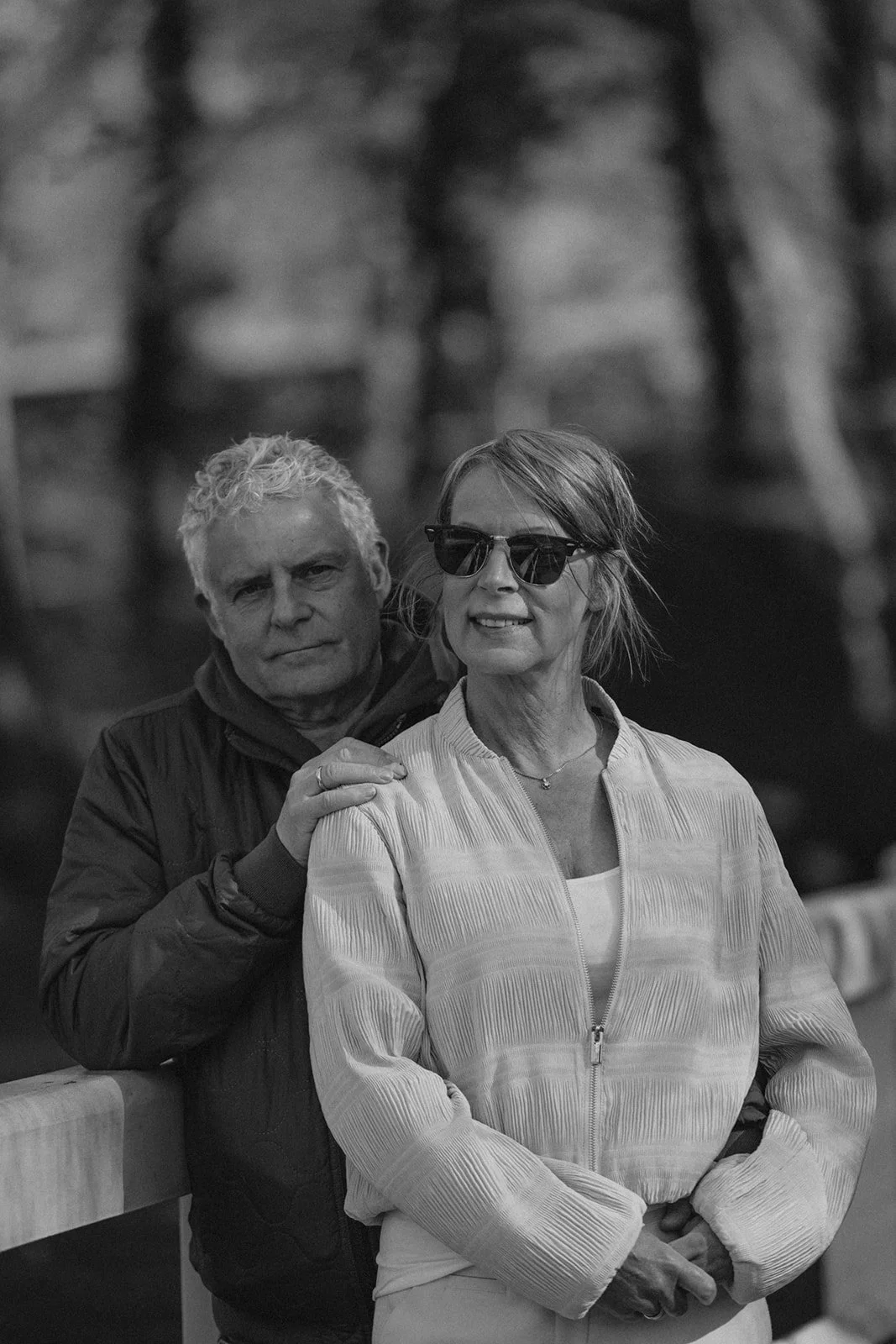 Ein älteres Paar steht an einem Geländer im Freien, der Mann hat weiße, lockige Haare und trägt eine dunkle Jacke; die Frau trägt Sonnenbrille, eine helle Jacke und hat kurze Haare. Das Bild ist in Schwarz-Weiß aufgenommen.