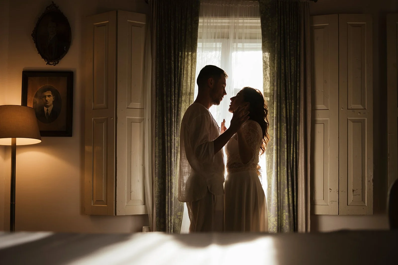 Ein Paar steht sich verliebt vor einem Fenster, im warmen Licht, in einem gemütlichen Raum mit alten Möbeln. Sie berühren sich zärtlich, es ist ein intimer Moment.