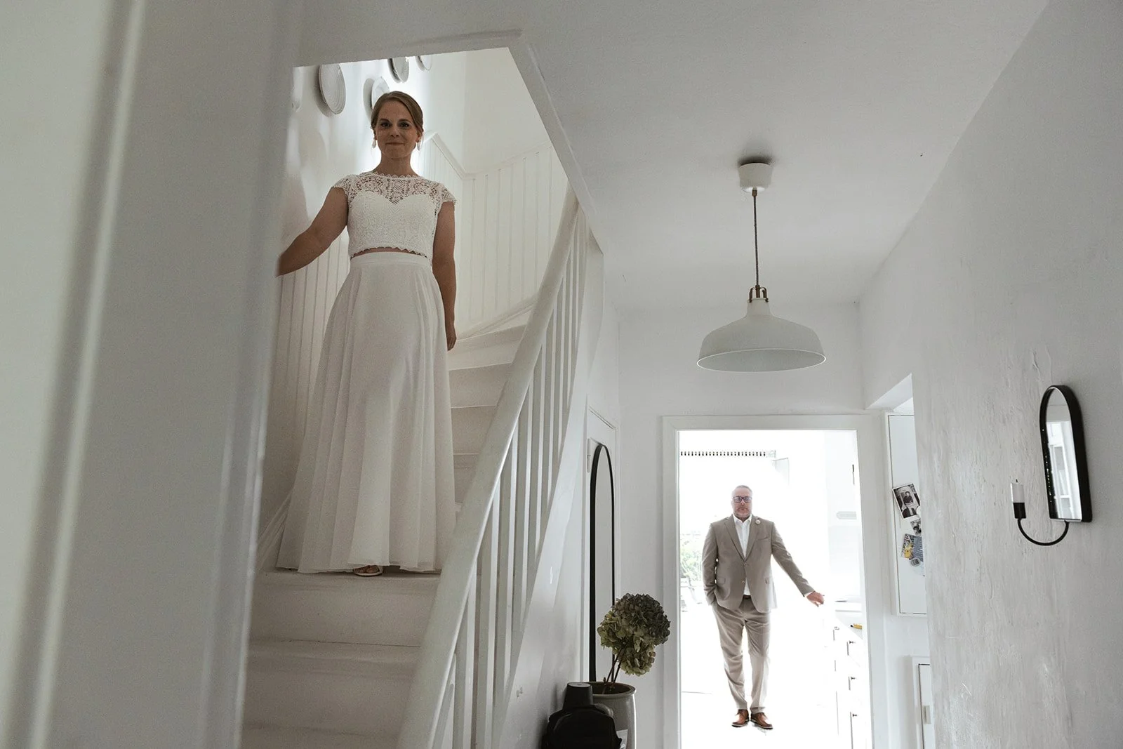 First Look Szene bei dem Paar zuhause. Der Raum ist zu sehen, an der Treppe steht die Braut, weiter hinten in der Küche wartet der Bräutigam. 
