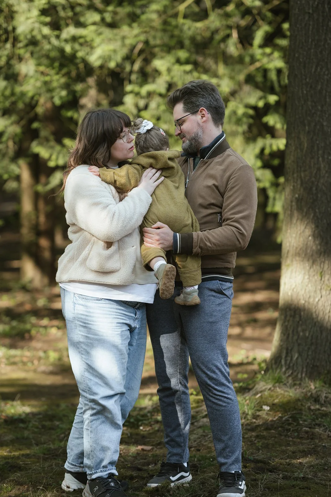 Familie aus zwei Erwachsenen und einem kleinen Kind, die sich im Park umarmen, umgeben von Bäumen und Natur.