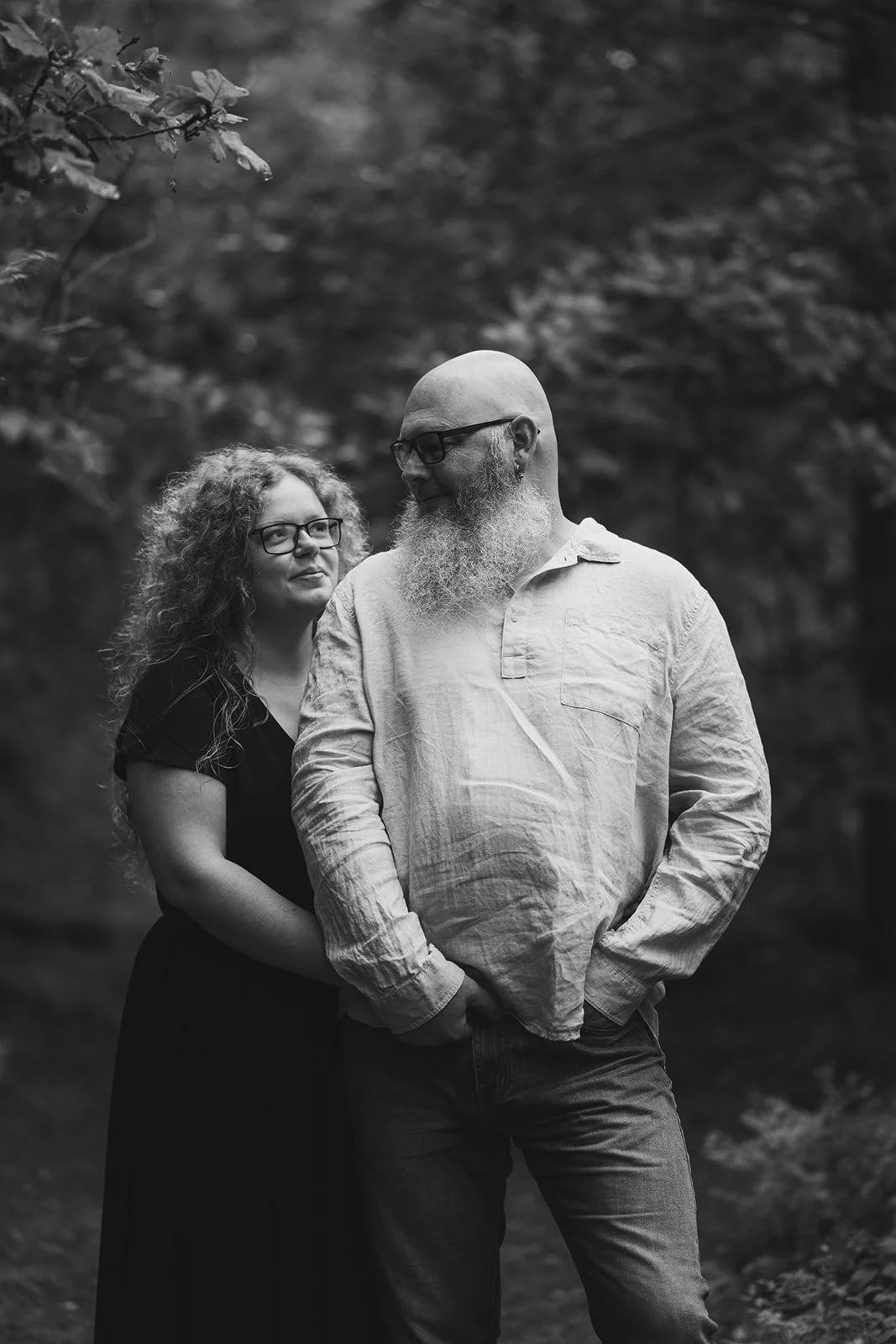 Schwarz-weiß-Foto eines Paares im Wald, die Frau steht hinter dem Mann, sie hält ihn an der Taille, beide tragen Brillen, die Frau hat lockiges Haar, der Mann hat einen Bart, sie schauen sich liebevoll an.