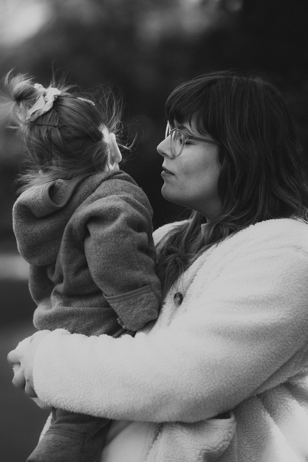 Die Mutter hält ein junges Mädchen im Arm. Sie stehen sich sehr nahe und schauen sich in die Augen, im Hintergrund ist ein unscharfer, natürlicher Hintergrund sichtbar. Das Bild ist schwarz-weiß.