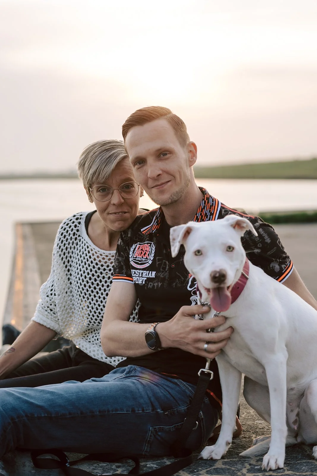 Ein Mann, eine Frau und ein Hund sitzen zusammen im Freien bei Sonnenuntergang am Wasser. Der Mann hält den weißen Hund mit braunem Fleck auf der Nase. Die Frau trägt Brille und ein weißes Netzoberteil. Der Mann trägt ein schwarzes T-Shirt mit bunten