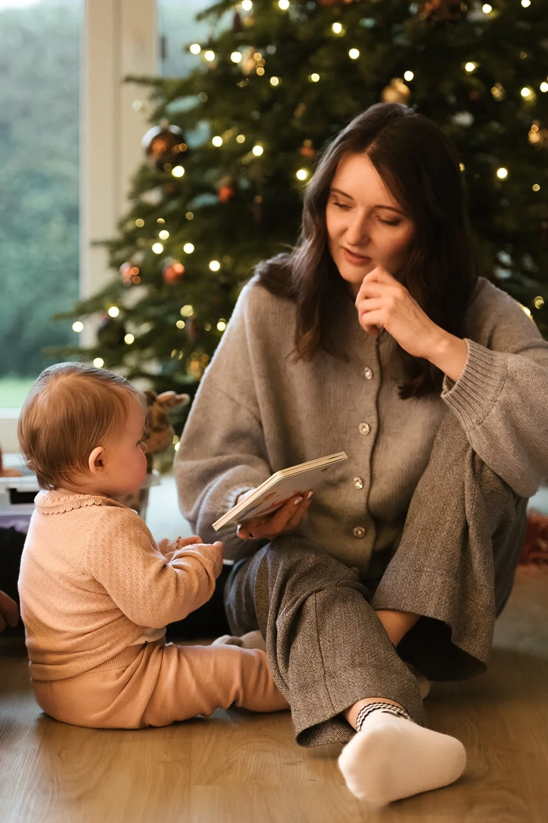 Eine Frau und ein kleines Kind sitzen vor einem Weihnachtsbaum, das Kind schaut die Frau an, während sie ein Buch liest.