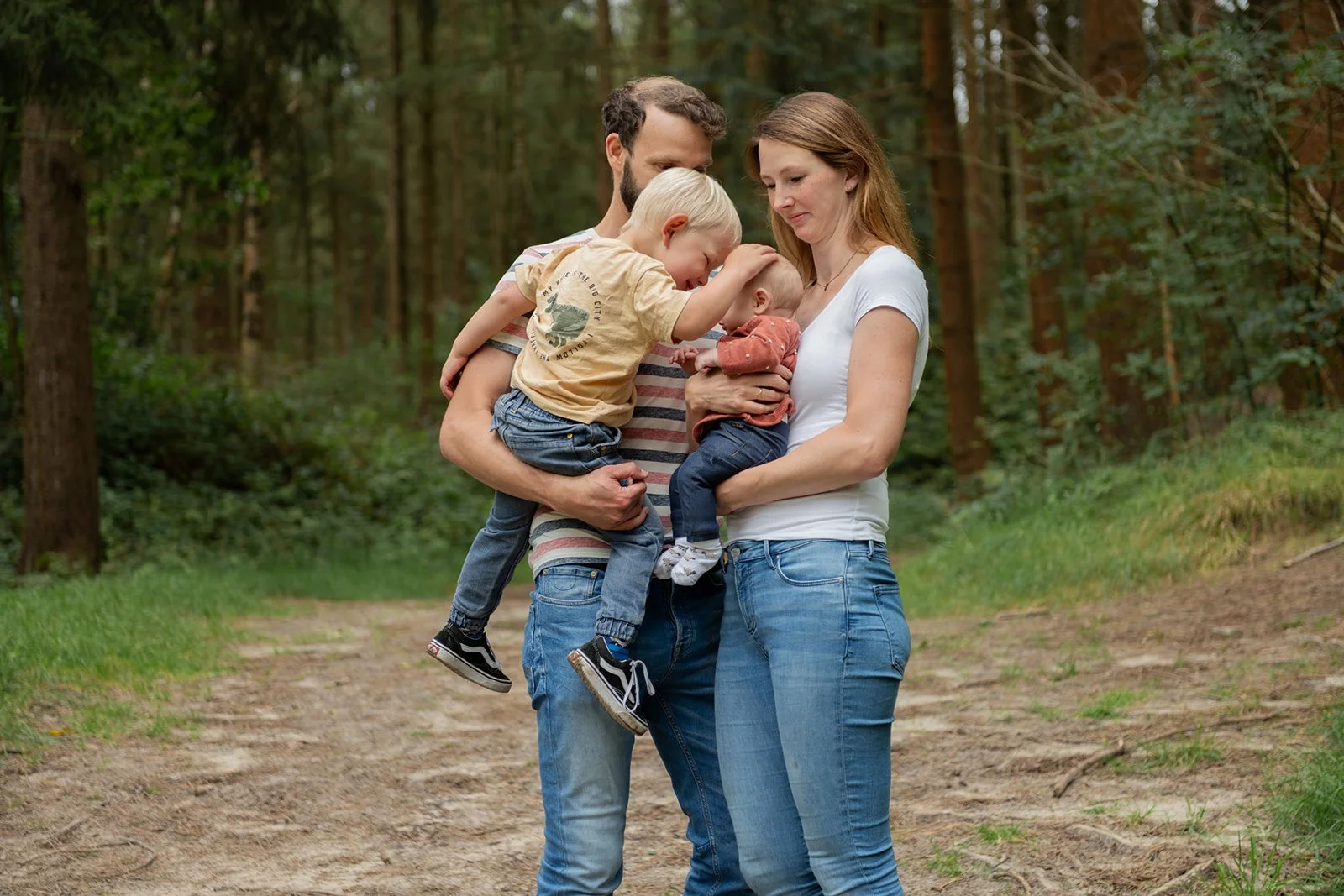 Familie mit zwei Kindern im Wald, Mutter und Vater halten die Kinder, während sie auf einem Waldweg stehen.