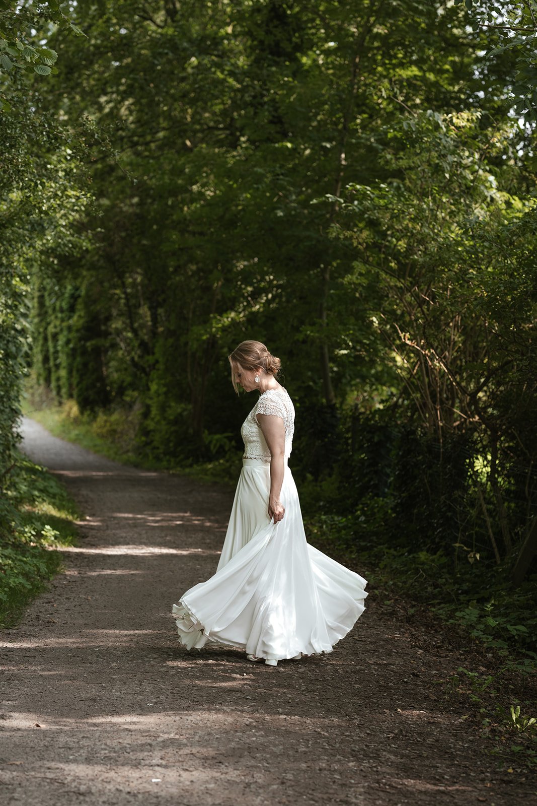 Eine Frau in einem weißen Brautkleid steht auf einem Waldweg, umgeben von grünen Bäumen.