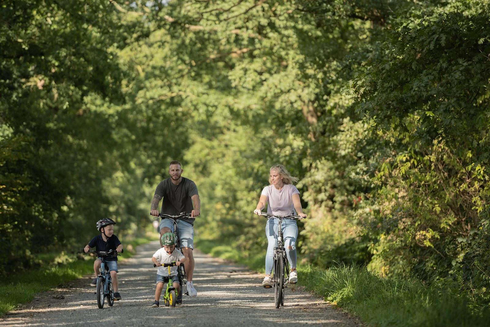 Familie mit zwei Erwachsenen und zwei Kindern beim Fahrradfahren auf einem Waldweg im Grünen.