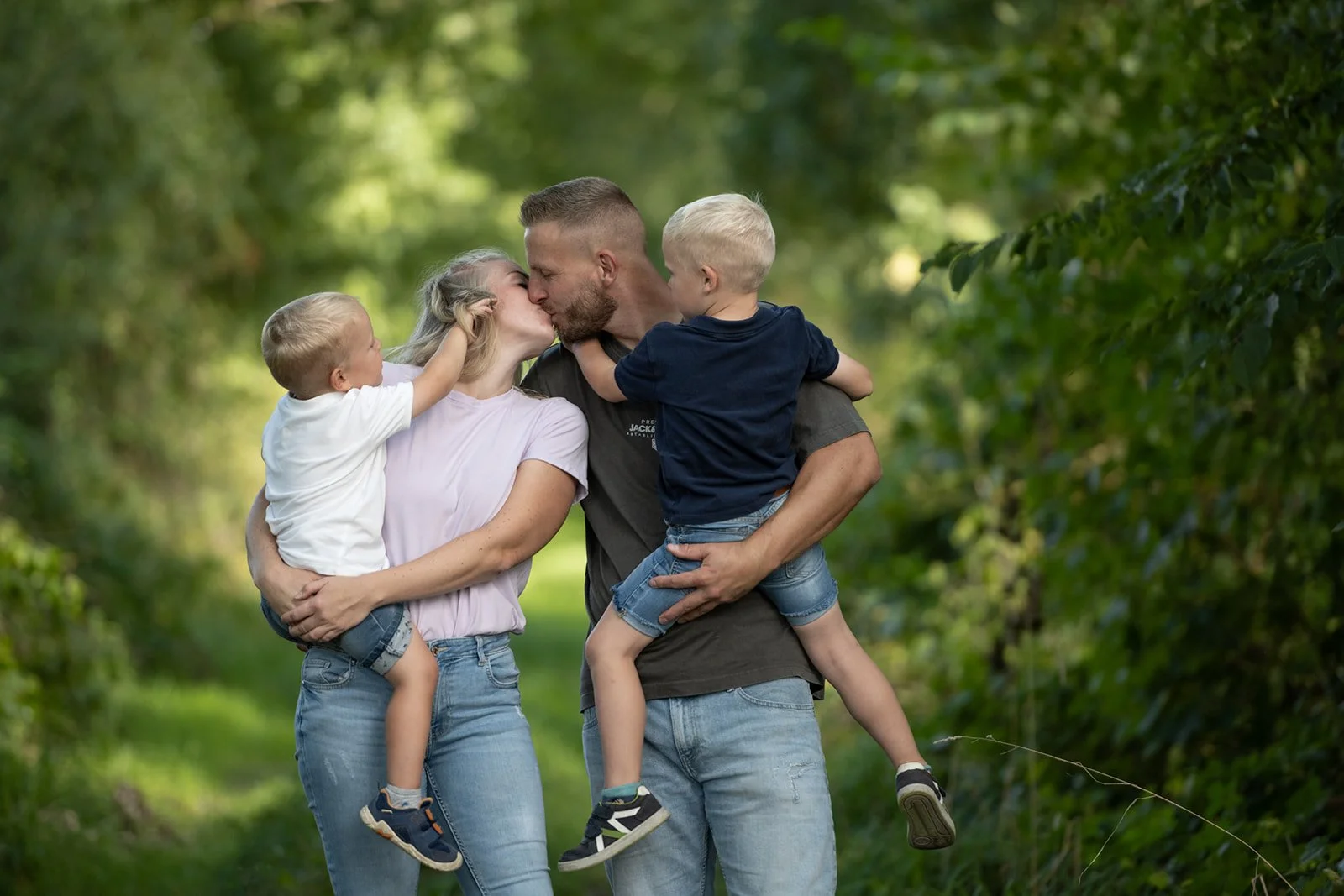Familie bei einem Spaziergang im Grünen. Die Eltern geben sich einen Kuss, während sie die Kinder im Arm halten.