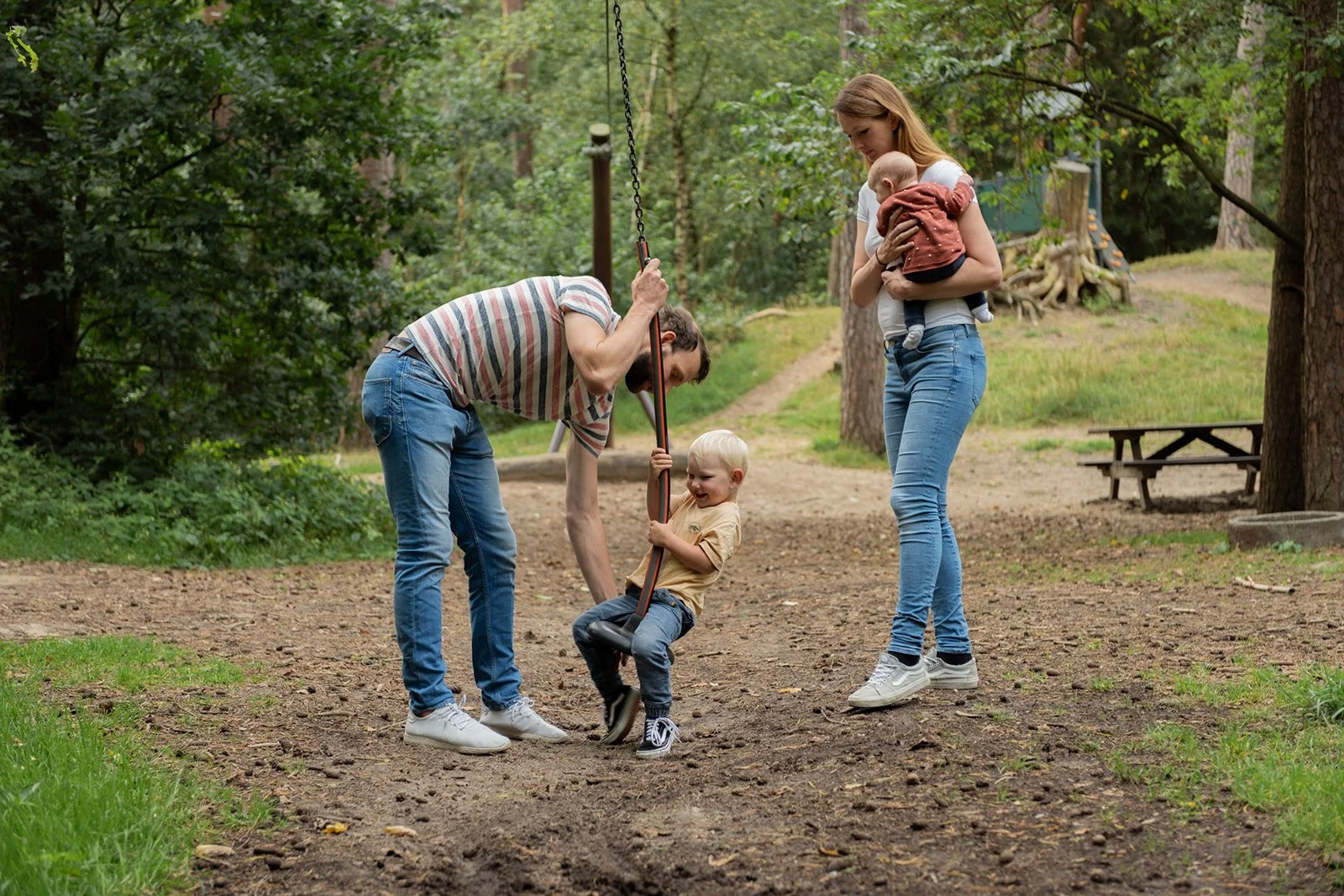 Familie mit einem kleinen Kind auf einer Schaukel im Park, während die Mutter ein Baby hält.