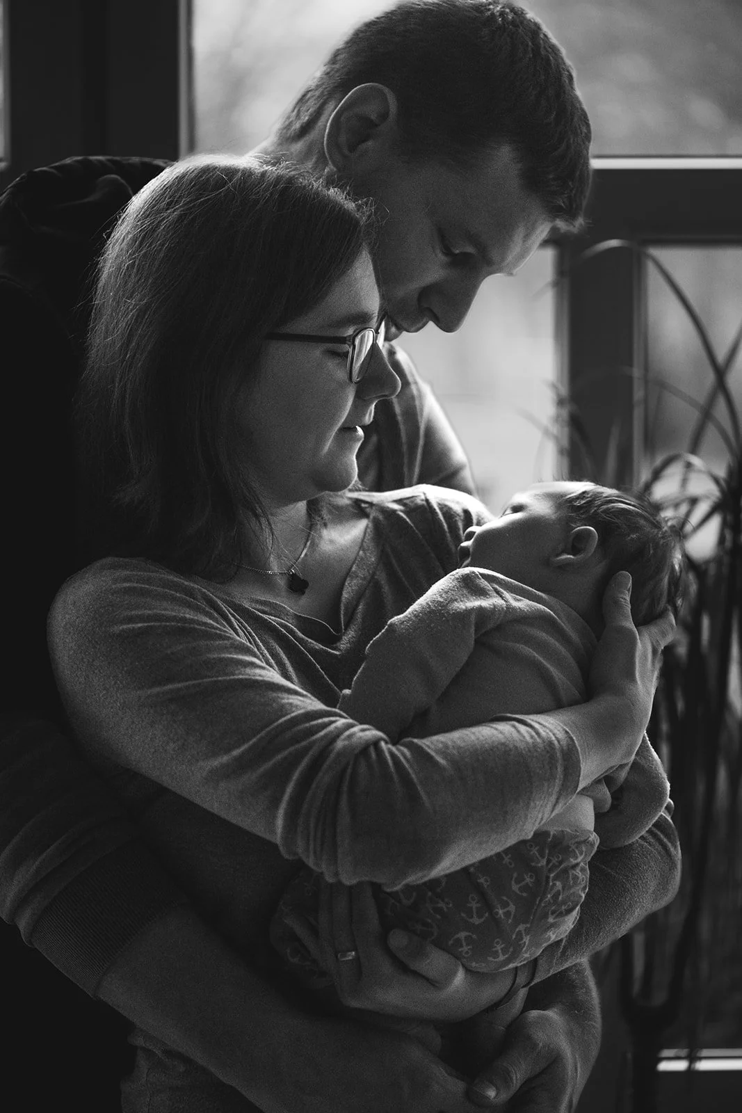 Eine Frau hält ein Baby im Arm, während ein Mann sich vorbeugt und die beiden schaut. Es ist eine Schwarz-Weiß-Fotografie, die eine intime und liebevolle Szene zeigt.