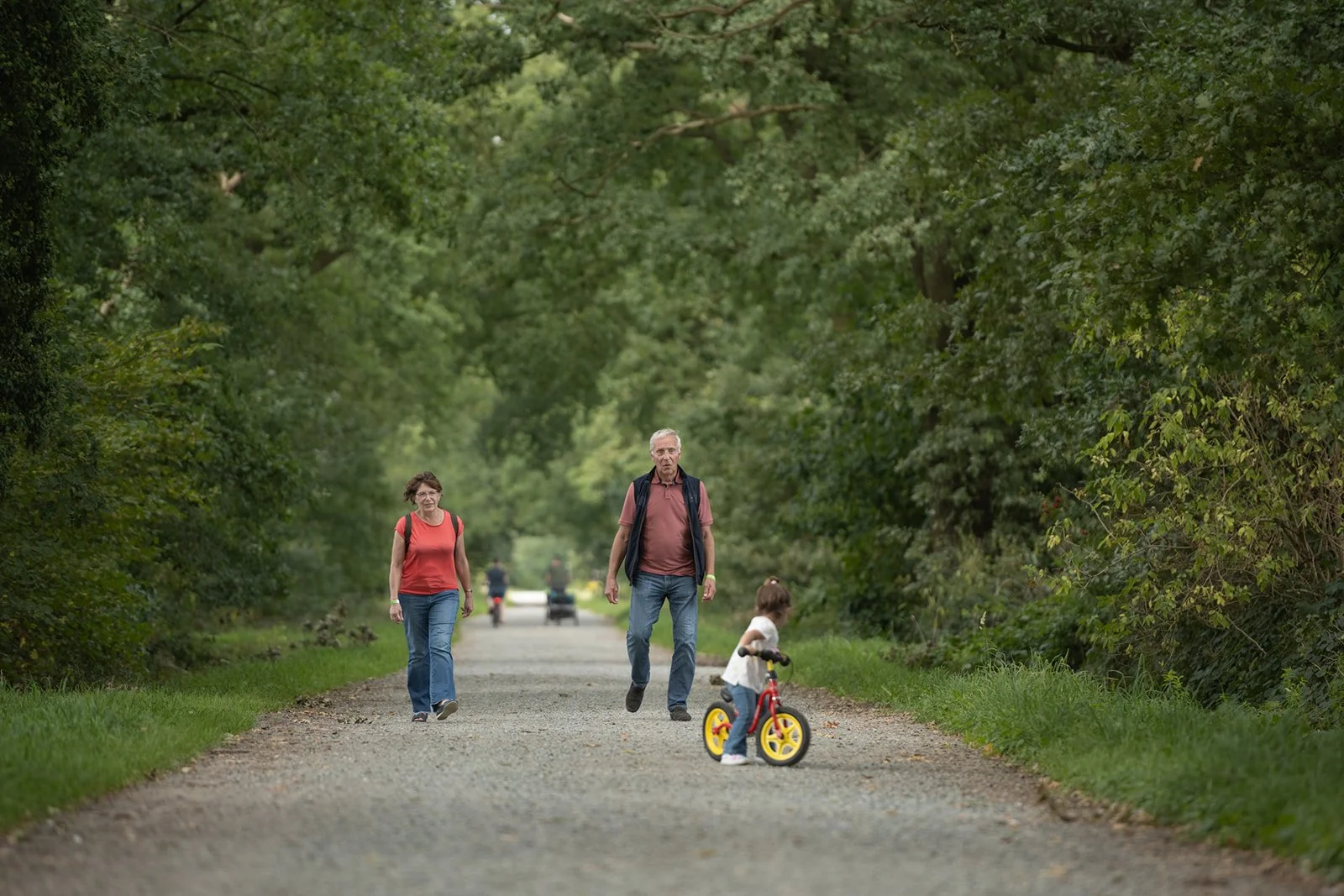 Eine Familie mit Kindern beim Spaziergang im Wald, ein Mädchen mit einem Fahrrad, zwei Erwachsene gehen auf einem schmalen Weg, umgeben von Bäumen.