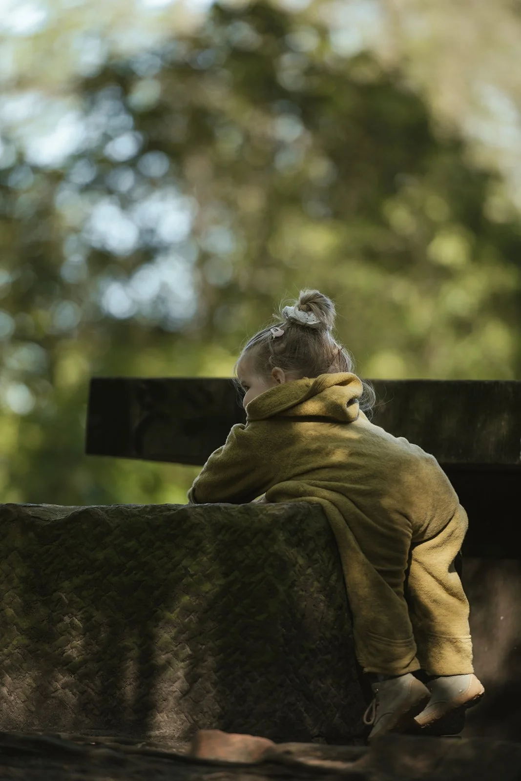 Ein Kind sitzt auf einem großen, wettergegerbten Baumstamm in einem Wald, trägt einen gelben Pullover und hat die Haare auf einem Zopf zusammengesteckt.
