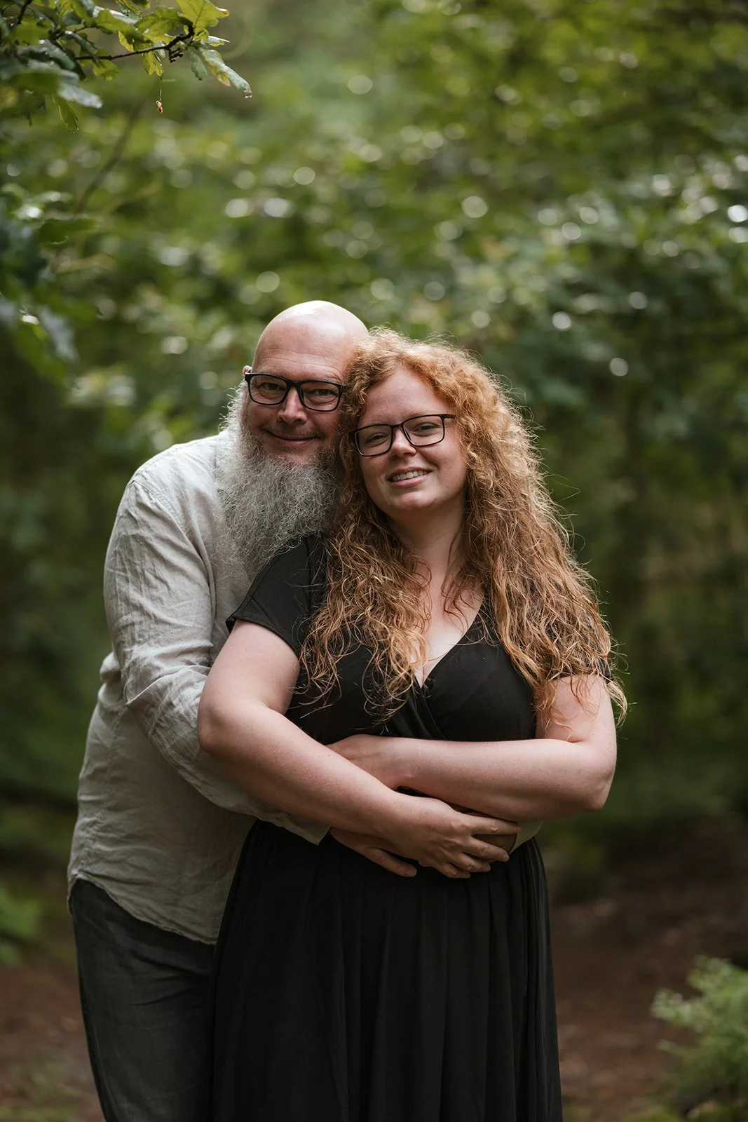 Ein Mann mit Brille und Bart umarmt eine Frau mit langen, lockigen roten Haaren und Brille in einem grünen Wald.