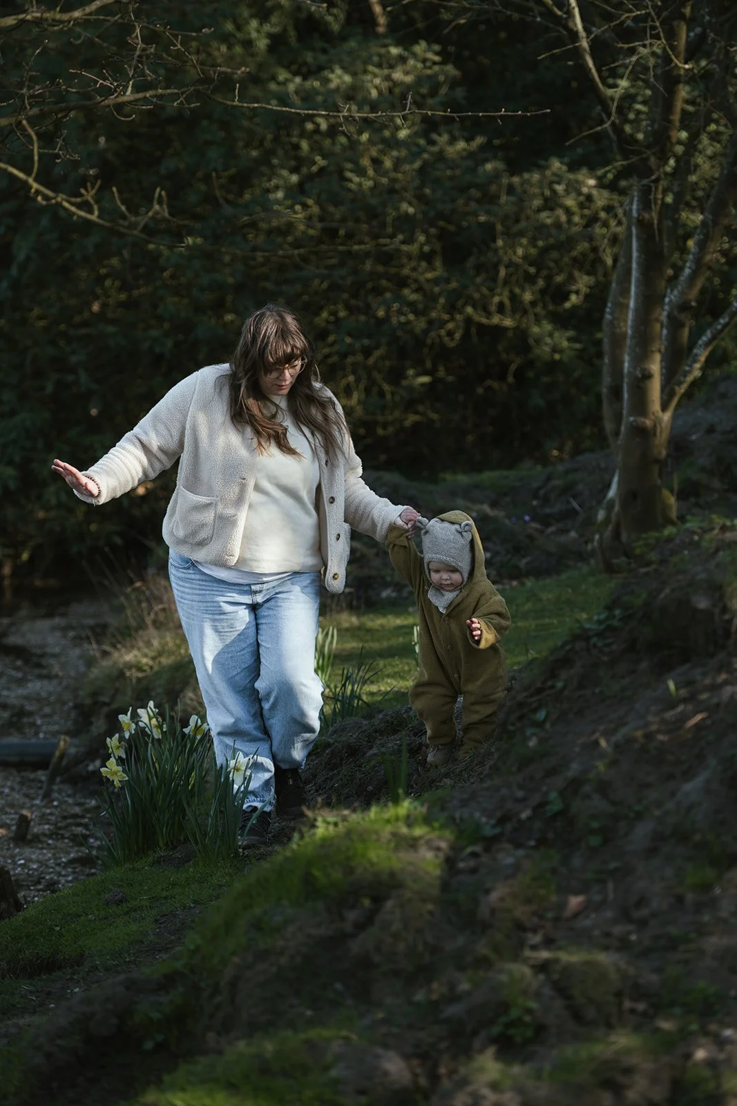 Eine Frau mit einem kleinen Kind beim Spaziergang im Park oder Wald. Die Frau trägt eine beige Jacke und blau Jeans, das Kind trägt einen braunen Anzug und eine graue Mütze. Sie gehen einen schrägen Weg entlang, umgeben von Büschen und Bäumen.