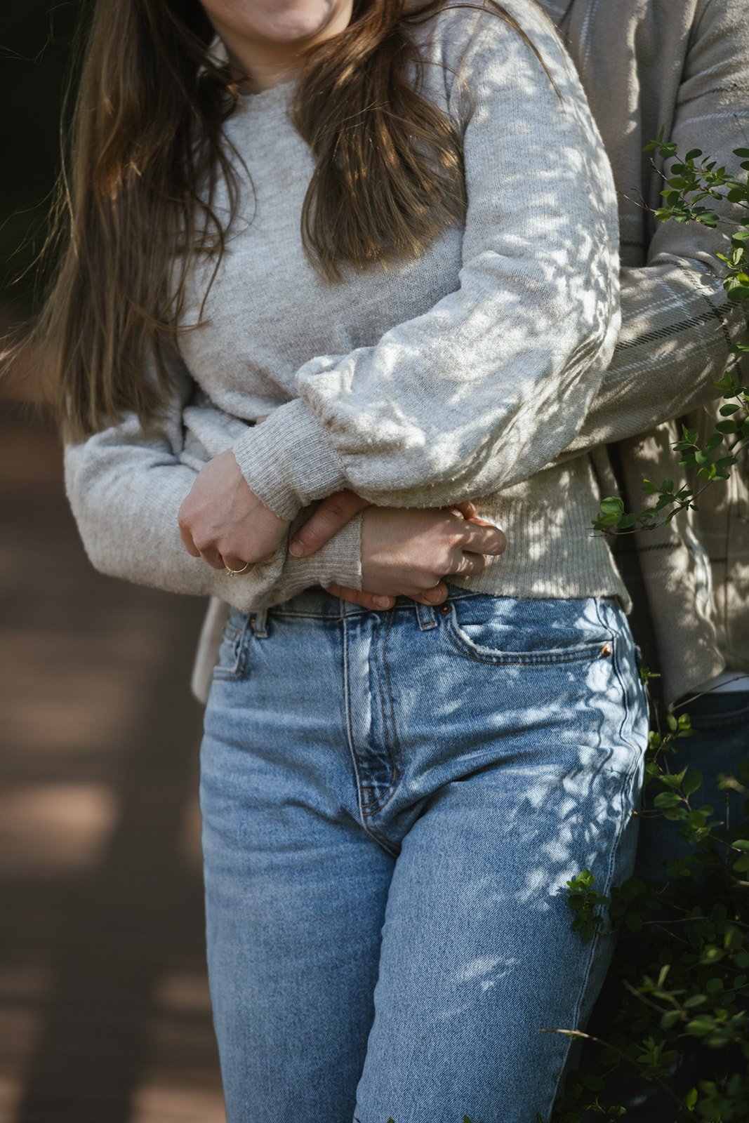 Nahaufnahme von zwei Person, die sich umarmen. die Frau trägt einen hellgrauen Pullover mit langen Ärmeln , Jeans. Hintergrund ist unscharf mit Schatten und Pflanzen.