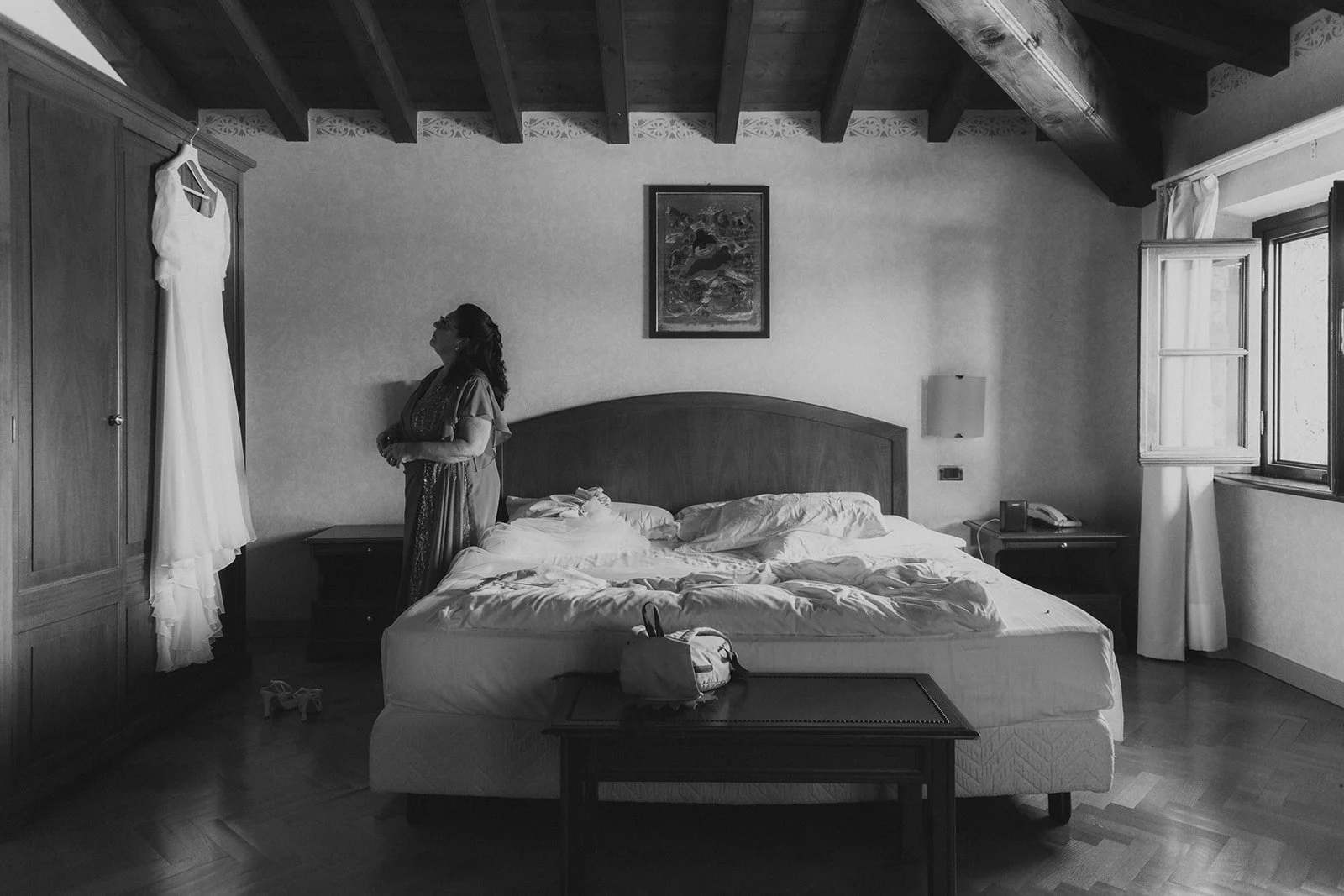 Frau steht in einem Hotelzimmer neben einem Bett, schaut auf ein weißes Hochzeitskleid, das an einem Kleiderbügel in einem Schrank hängt, das Zimmer ist aufgeräumt, das Bett ist unordentlich, es gibt eine Tagesdecke, Kissen, ein Nachttisch mit einem Telefon und eine Lampe, ein Bild an der Wand und ein Fenster mit geöffneten Jalousien.