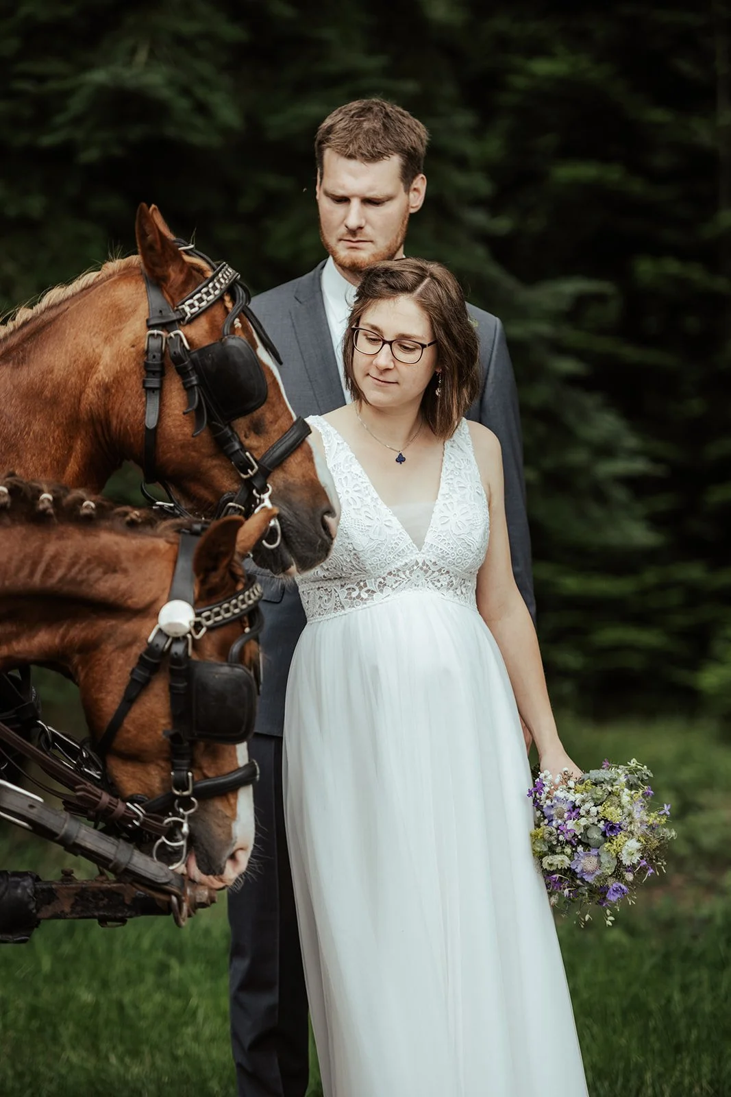 Ein Braut und Bräutigam stehen im Grünen, die Braut hält einen Blumenstrauß in der Hand, während sie ein braunes Pferd betrachten.