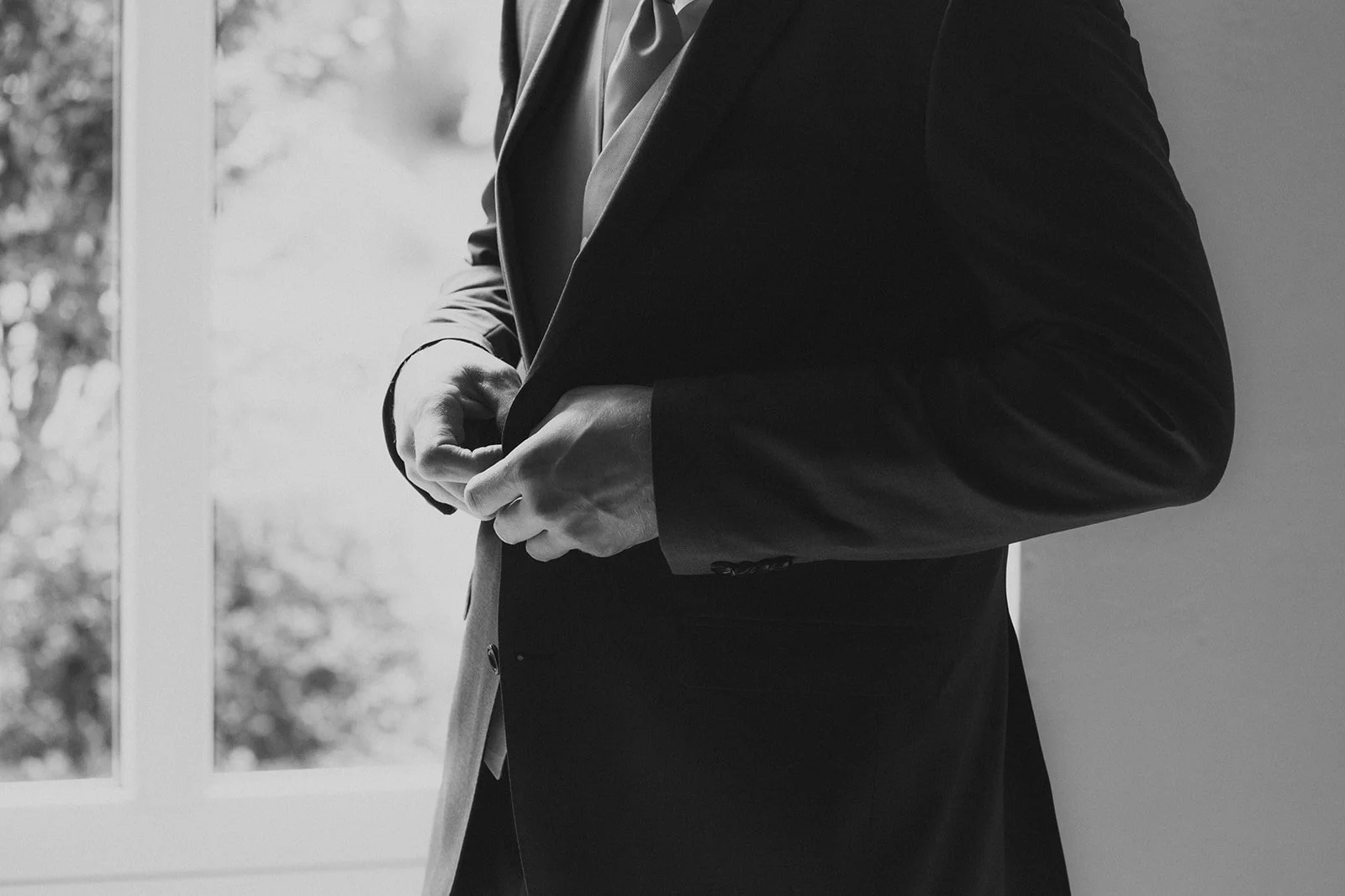 Ein Mann in einem Anzug ist dabei, seine Jacke zuzuknöpfen, neben einem Fenster mit Blick auf Bäume.