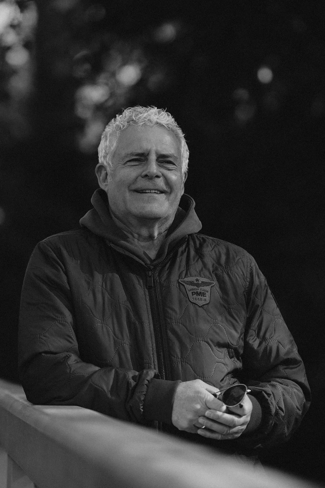 Ein älterer Mann mit kurzen, weißen Haaren und einem Lächeln, der eine dunkle, wattierte Jacke trägt, auf der ein Abzeichen mit der Aufschrift "PME" sichtbar ist.