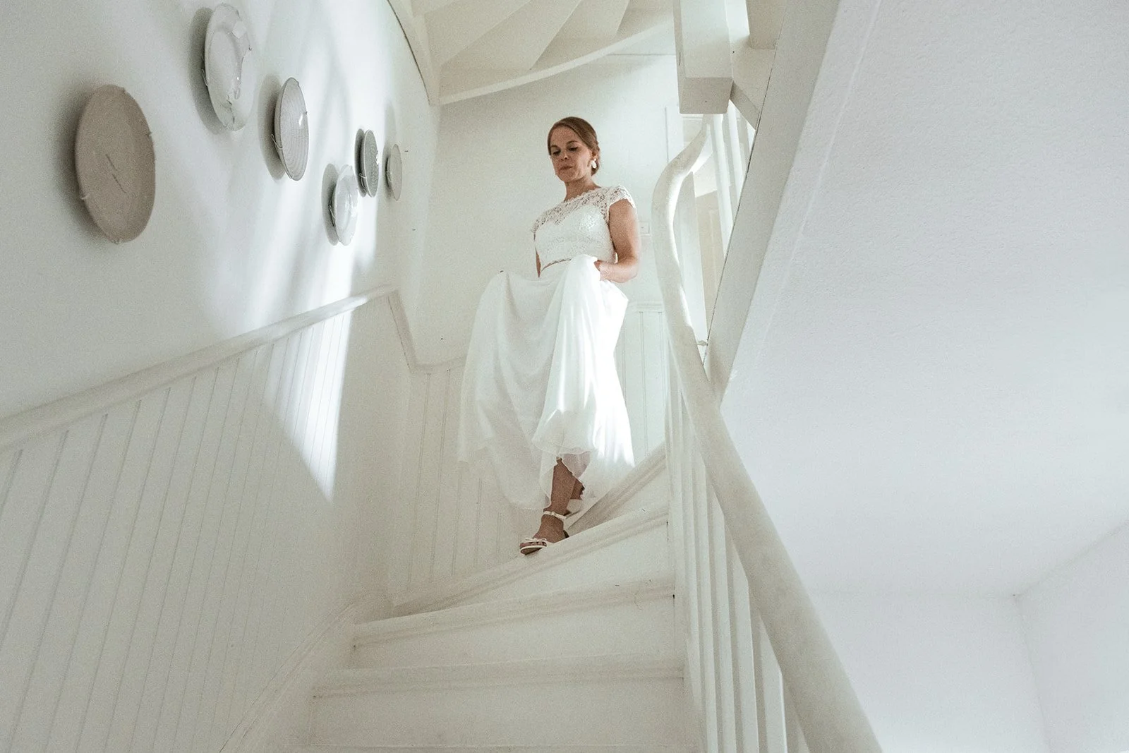 Frau in weißem Hochzeitskleid steht auf einer weißen Treppe im Haus.