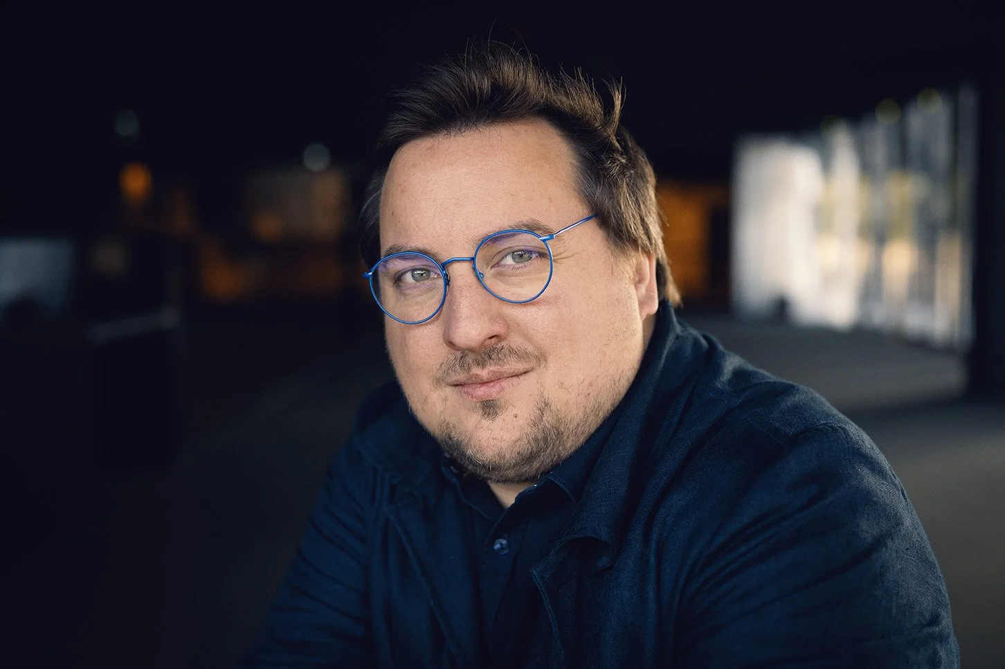 Portrait d'un homme - Adrien ramon - avec des lunettes rondes bleues, barbe et cheveux bruns, portant une chemise ou un manteau bleu foncé, dans un lieu intérieur avec une lumière naturelle et floue en arrière-plan.