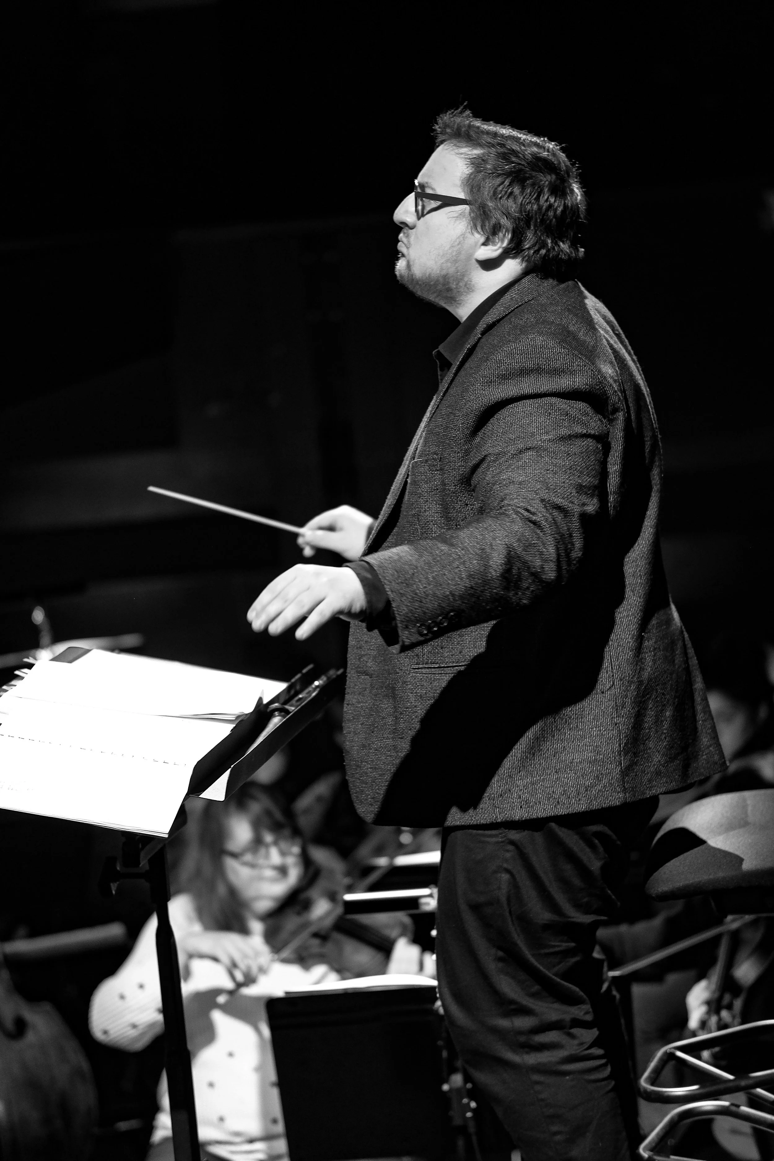 Un chef d'orchestre homme portant des lunettes, en costume, dirigeant un orchestre dans une performance, photo en noir et blanc.