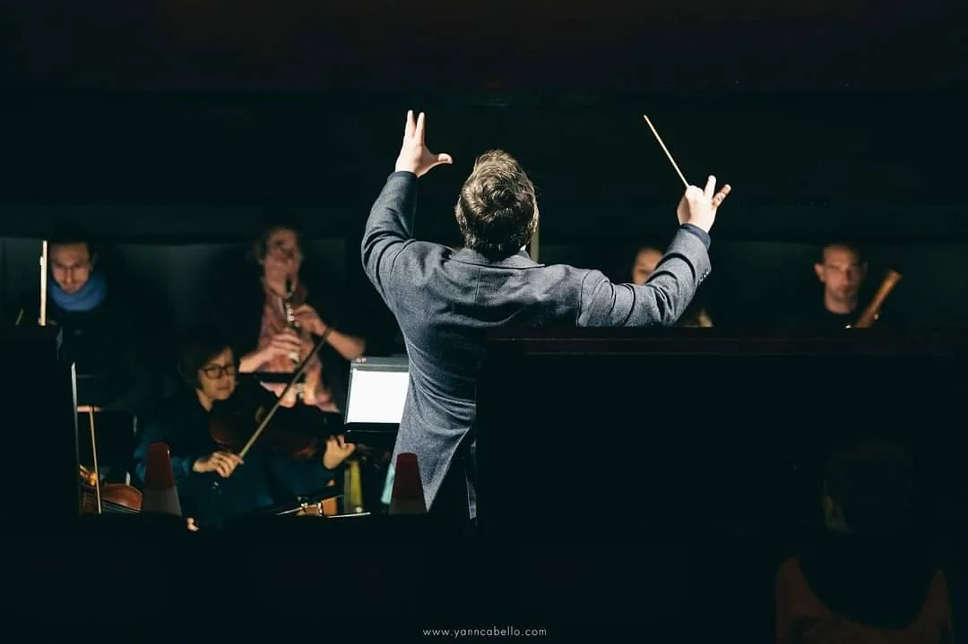 Chef d'orchestre dirigeant un ensemble musical dans une salle sombre.