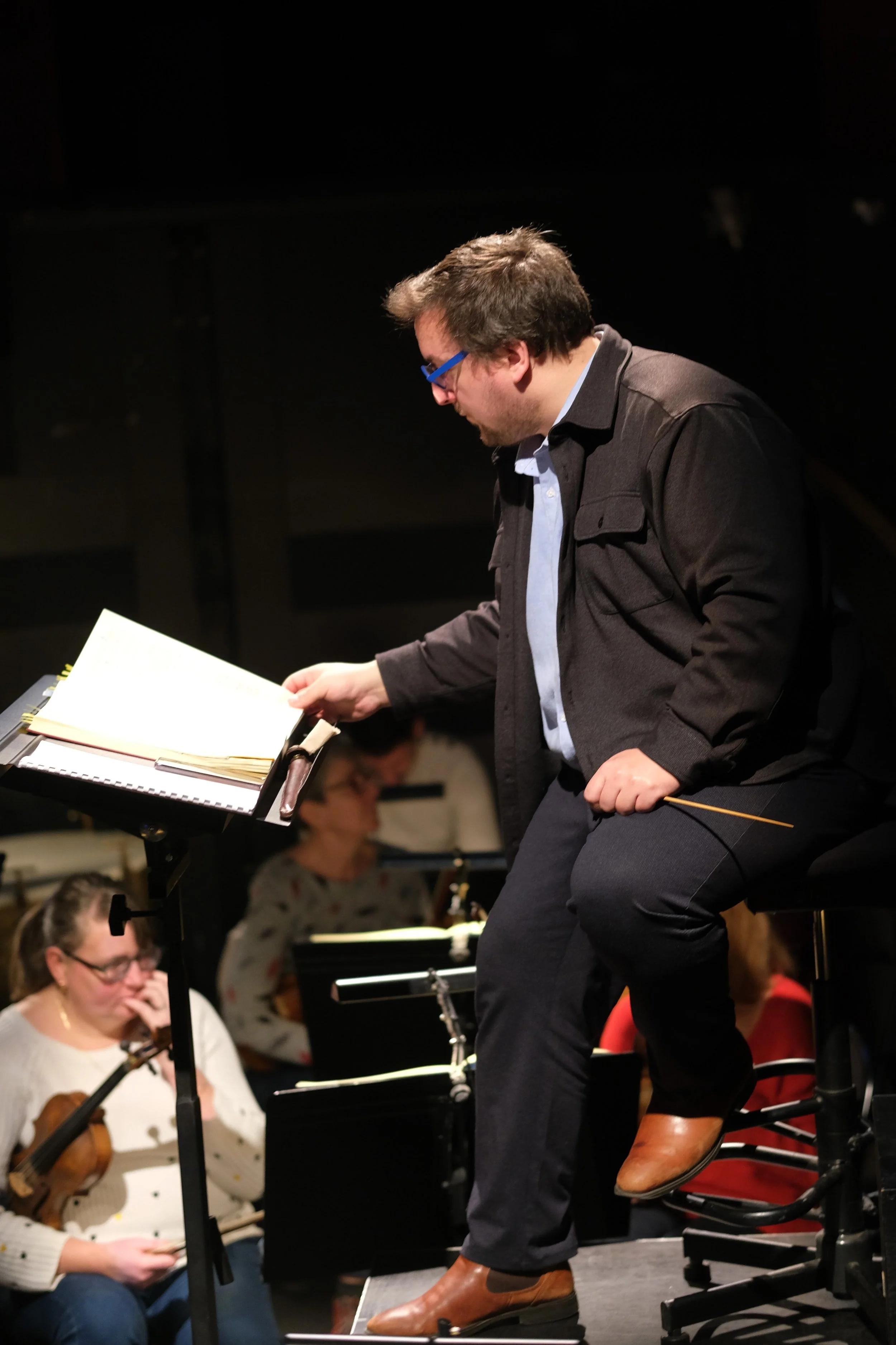 Un homme avec lunettes bleues, en costume noir, assis sur un tabouret, tient une baguette de chef d'orchestre. En arrière-plan, on peut voir quelques musiciens avec des instruments, dont une femme jouant du violon, en train de préparer ou jouer dans un orchestre.