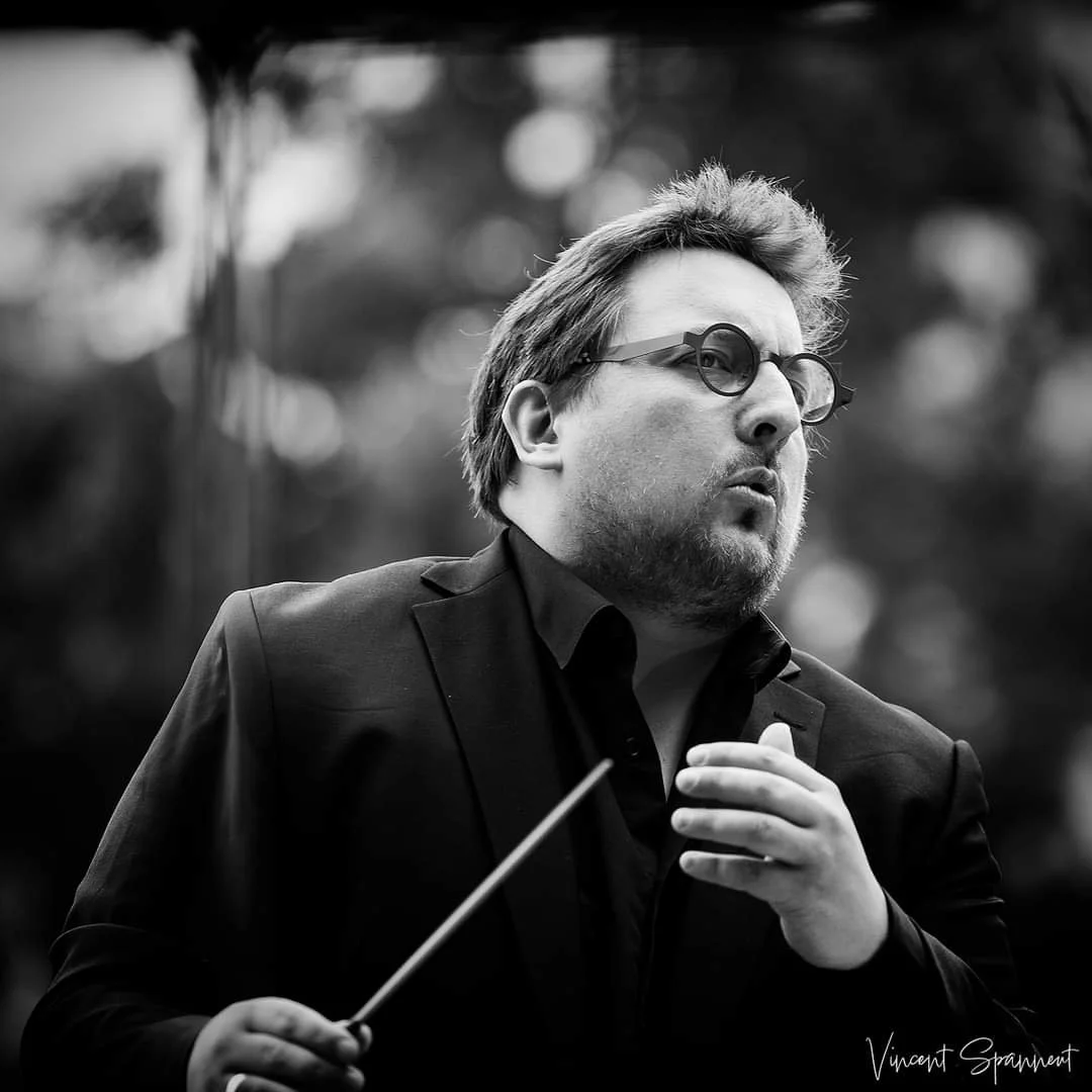 Un homme en noir et blanc portant des lunettes rondes, dirigeant un orchestre ou un chœur, tenant un bâton de direction, dans un geste concentré.
