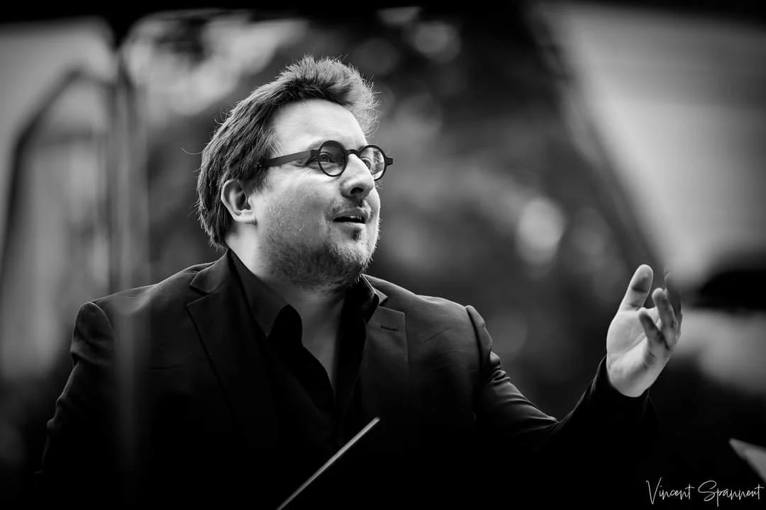 Un homme avec des lunettes, portant un costume, parle ou chante, avec un fond flou.