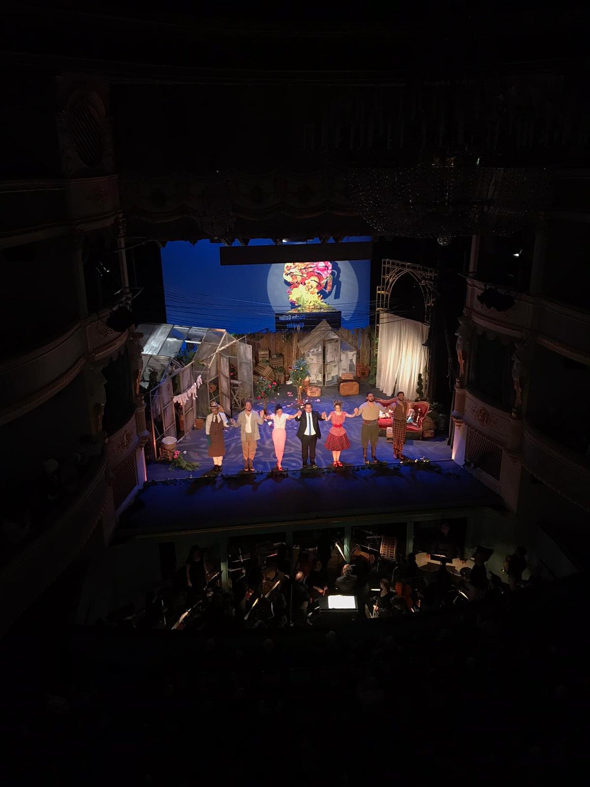 Acteurs sur scène dans un théâtre, après une représentation, en train de saluer le public, avec un orchestre en dessous, avec un décor de maison et une projection d'image colorée en fond.