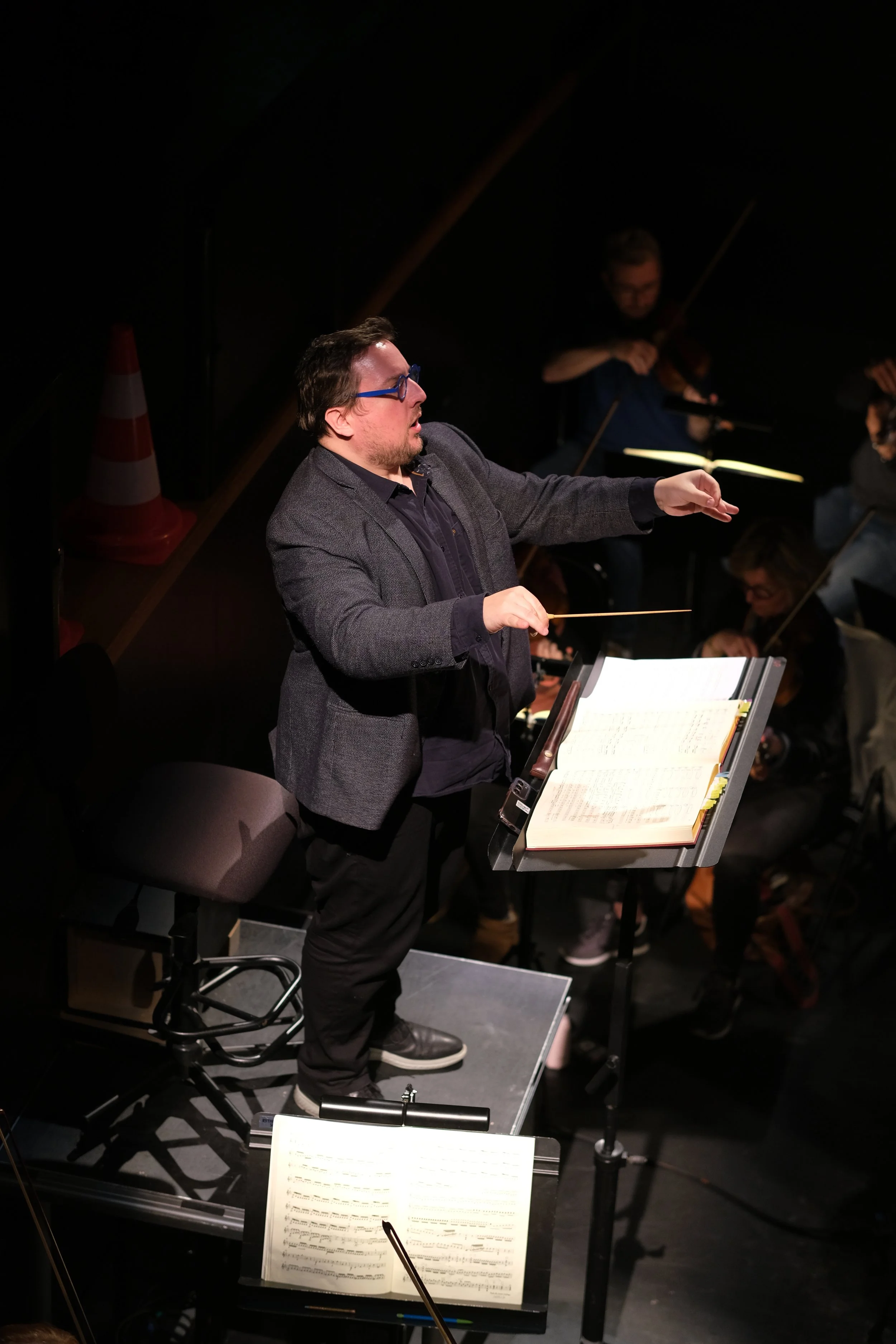 Un chef d'orchestre - Adrien ramon - en costume gris, avec des lunettes bleues, conduisant un orchestre lors d'une performance, avec des musiciens en arrière-plan.