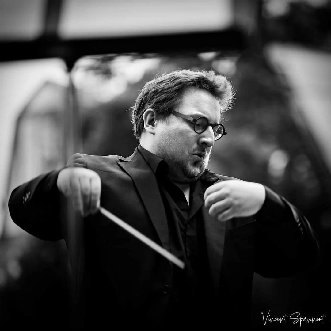 Un homme avec des cheveux courts, portant des lunettes rondes noires et un costume sombre, tient un bâton de dirigeant d'orchestre, fixant intensément. Il est en pleine direction, concentré et expressif.