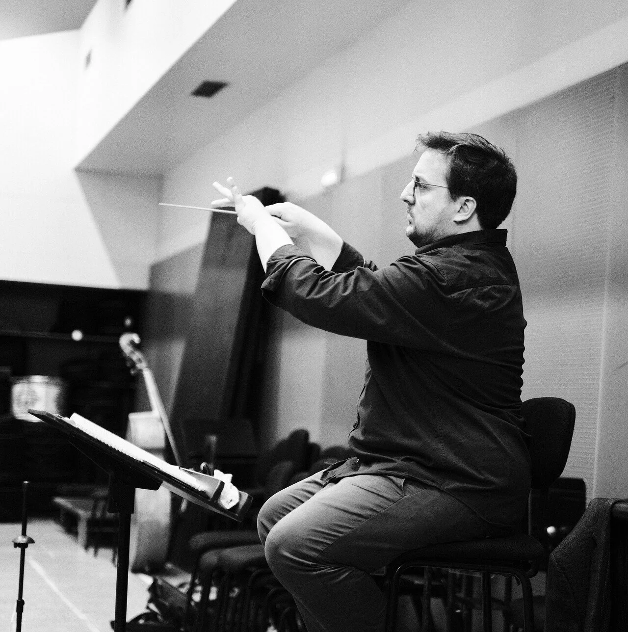 Un homme - Adrien ramon - en train de diriger avec un bâton dans une salle de concert, assis sur une chaise, en noir et blanc.