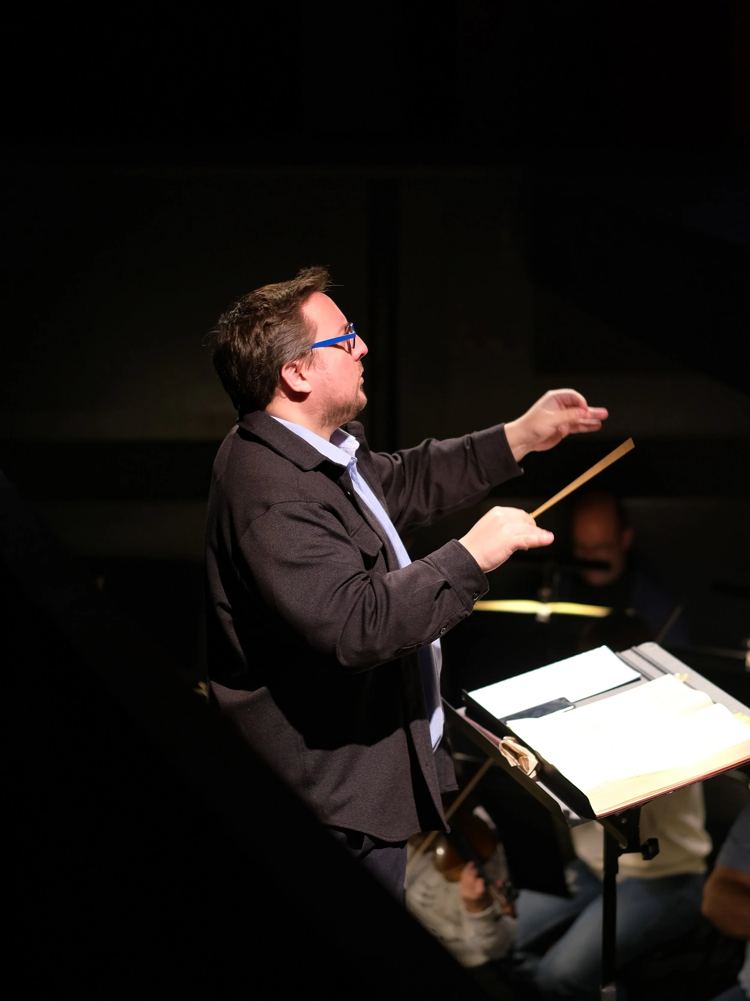 Un homme dirigeant un orchestre avec une baguette, placé devant une partition, dans un environnement sombre.