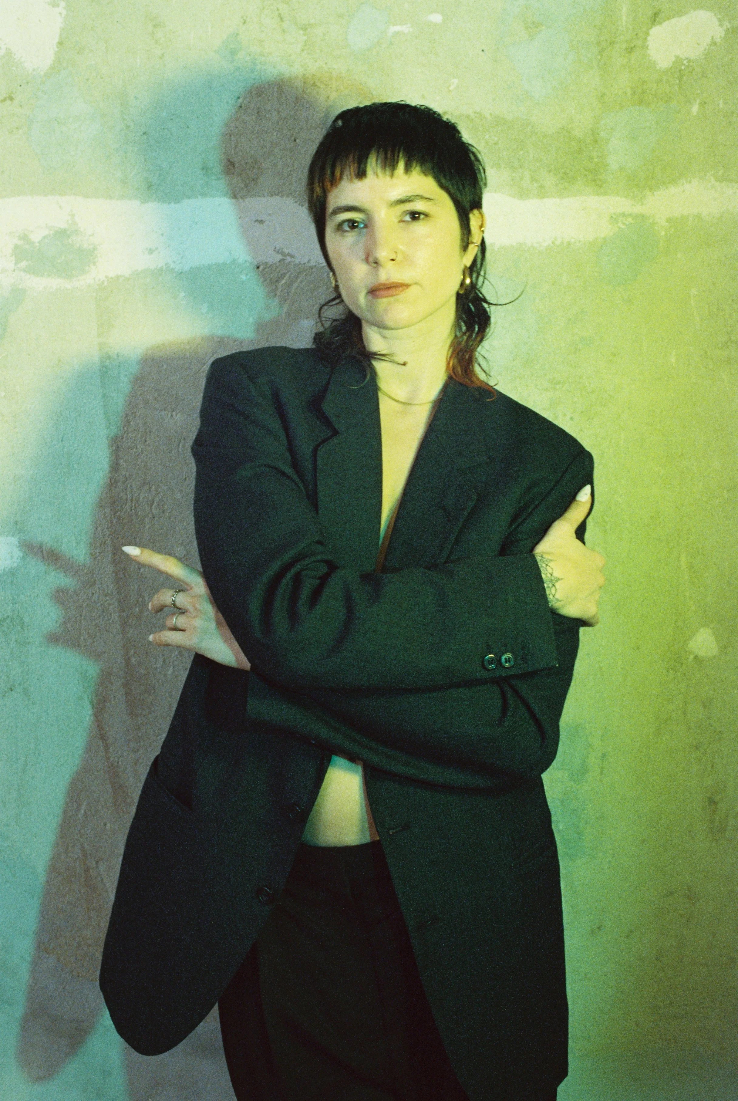 The author Mars de Beaucourt with dark, short hair and pale skin wearing a black blazer stands against a textured wall with colored lighting, crossing their arms and looking at the camera.