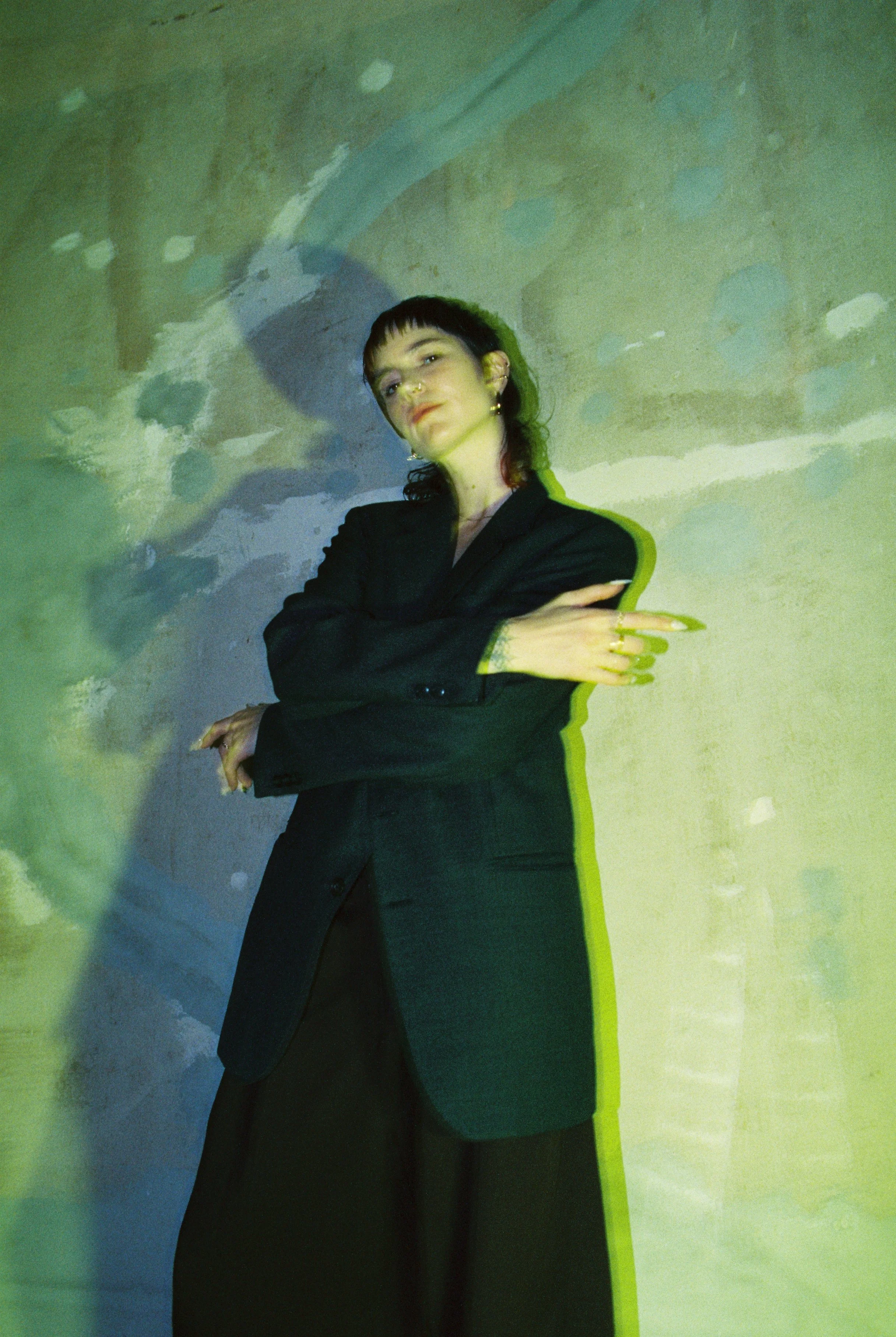 The author Mars de Beaucourt stands against a textured wall with green and yellow lighting effects, wearing a black blazer and making a crossed-arm gesture.