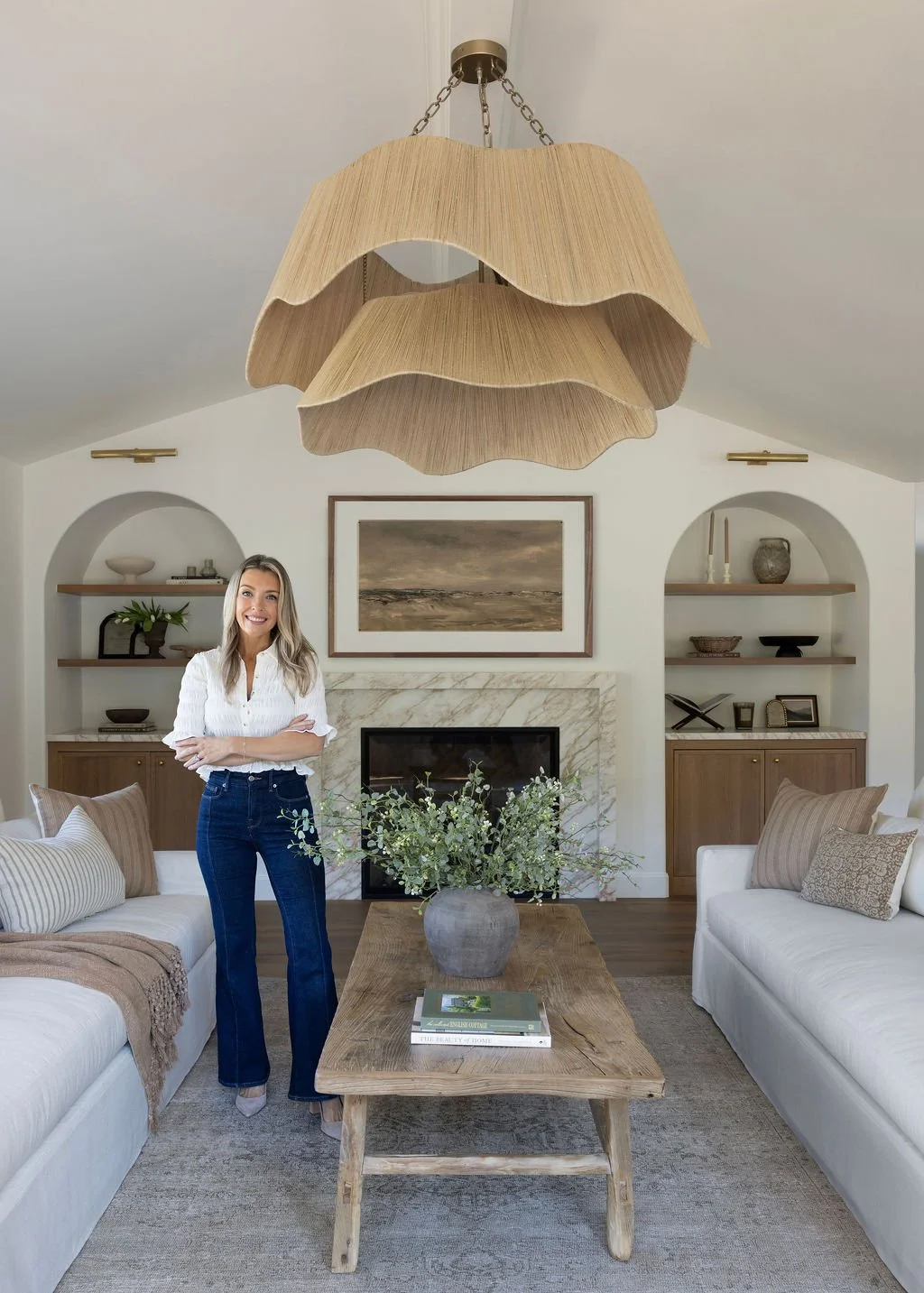 A woman standing in a well-decorated living room with a large wooden coffee table, a fireplace, built-in shelves, and a unique layered fabric lampshade hanging from the ceiling.