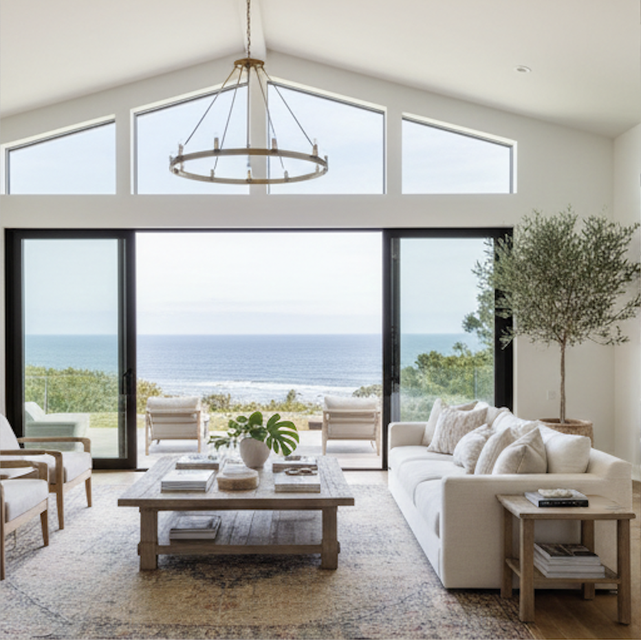 Living room with large glass sliding doors opening to ocean view, with a beige sofa, wooden armchairs, a wooden coffee table, a floor rug, and potted plants under a high ceiling with triangular windows and a chandelier.
