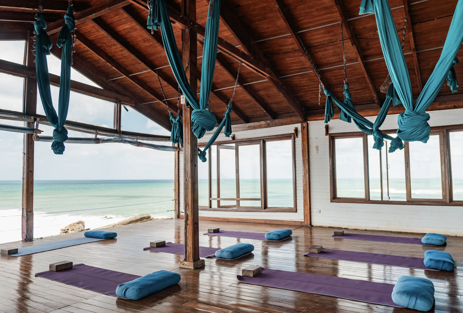 A yoga studio with purple mats and blue bolsters arranged on a wooden floor. Blue silk aerial yoga hammocks hang from the ceiling, and large windows reveal an ocean view outside.