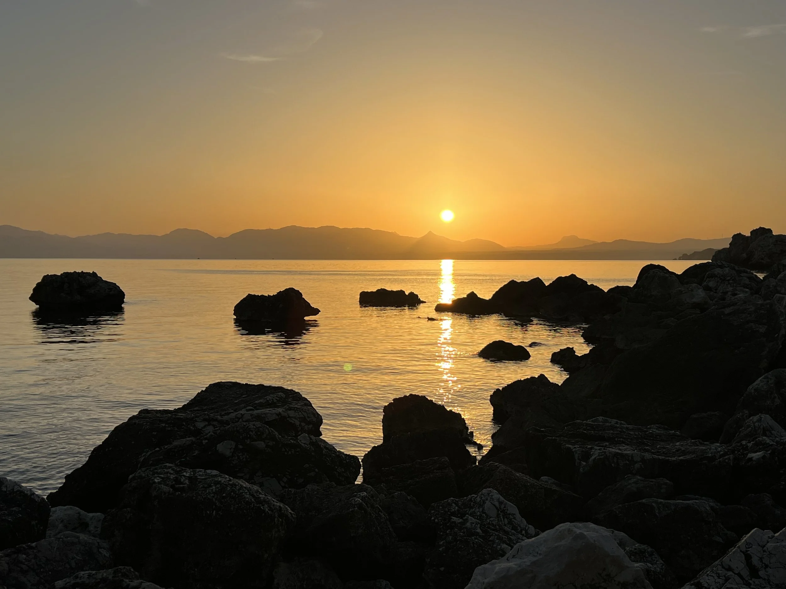 A sunset over a body of water with mountains in the background and rocks along the shoreline in the foreground
