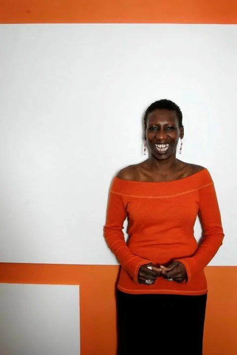 Smiling woman in an orange off-shoulder sweater standing against a white and orange wall.