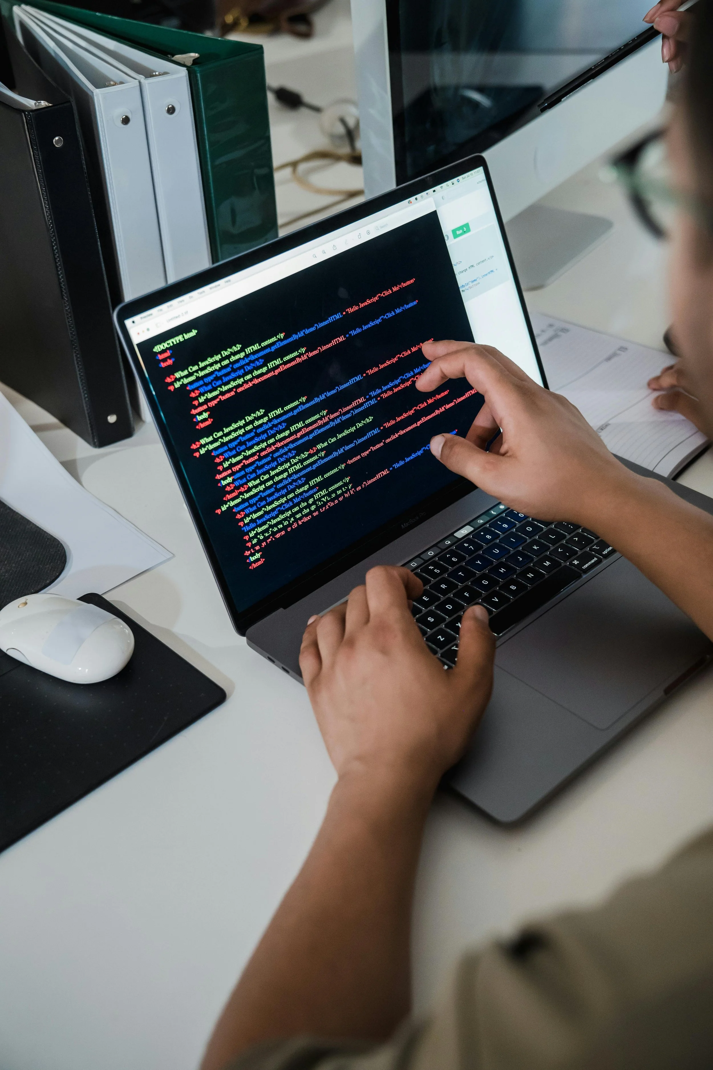 Person programming on a MacBook with colorful code on the screen, surrounded by office supplies and a computer monitor.