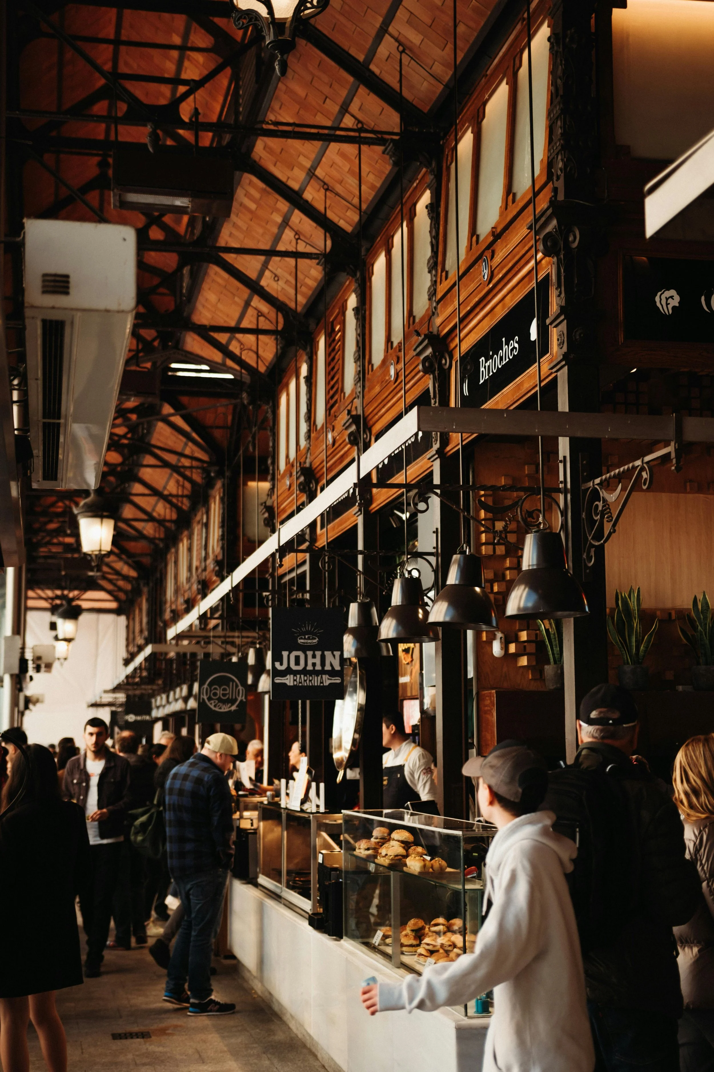 A busy indoor food market with people lining up at food stalls, featuring menu signs, and baked goods displayed in a glass case.