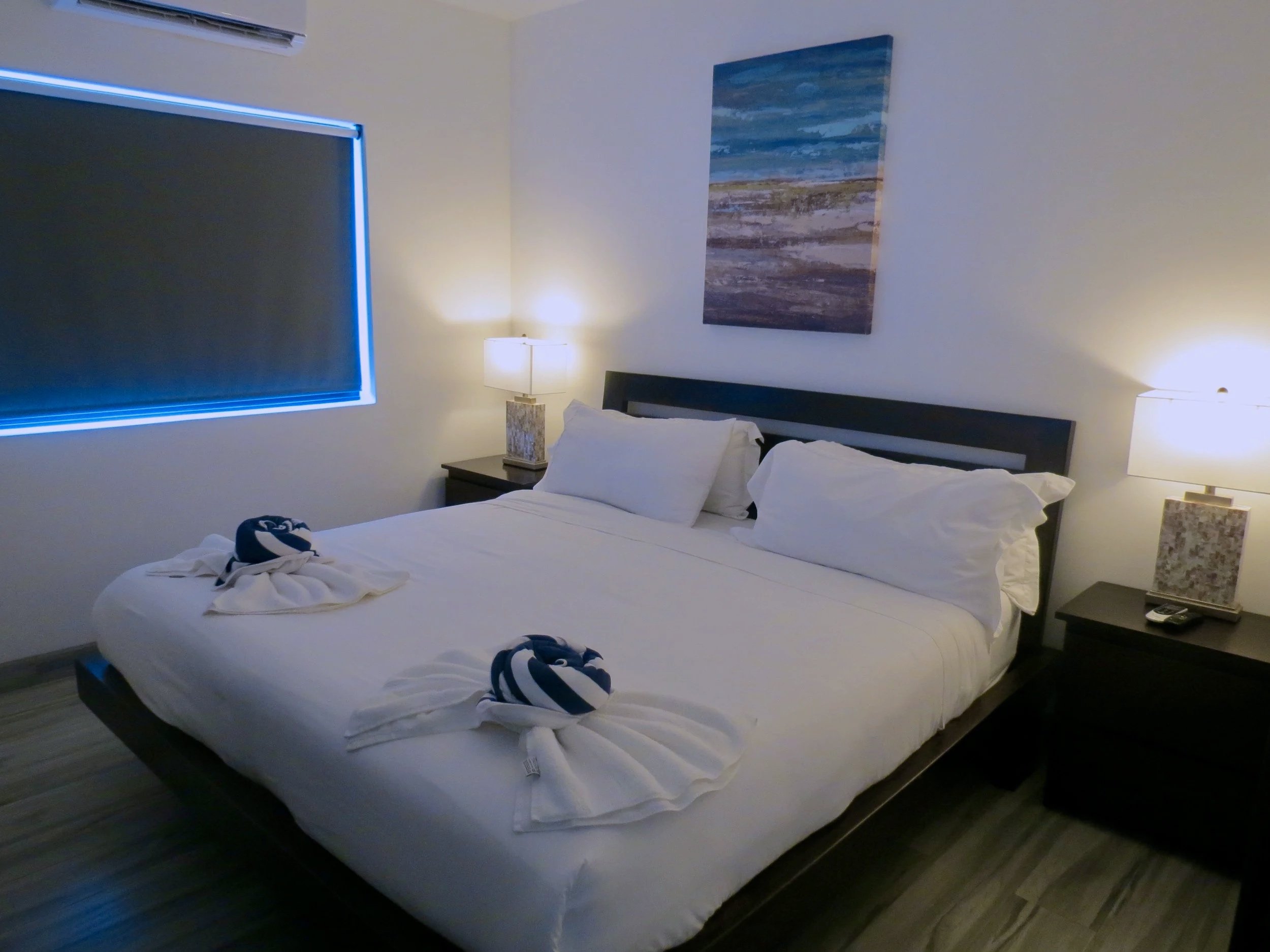 Guest bedroom with a neatly made bed, two bedside tables with lamps, and a painting of a beach scene on the wall. The window has a blackout blind.  Unit S2, Caye Caulker,  Blu Zen Belize