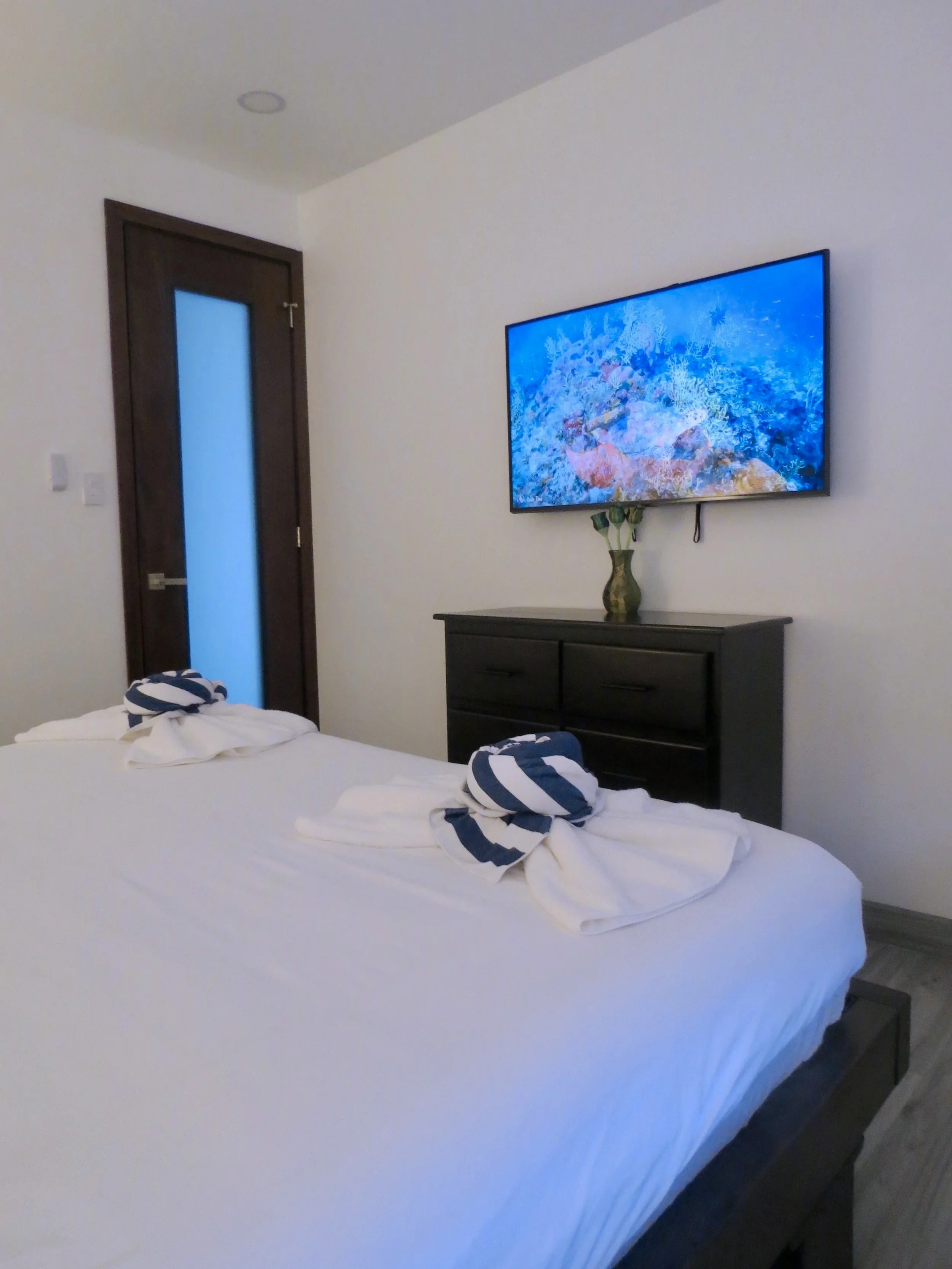 Guest bedroom with a king-size bed, towels folded with striped fabric accents, a dresser with a vase, and a wall-mounted television showing an underwater scene.  Unit S2, Caye Caulker,  Blu Zen Belize