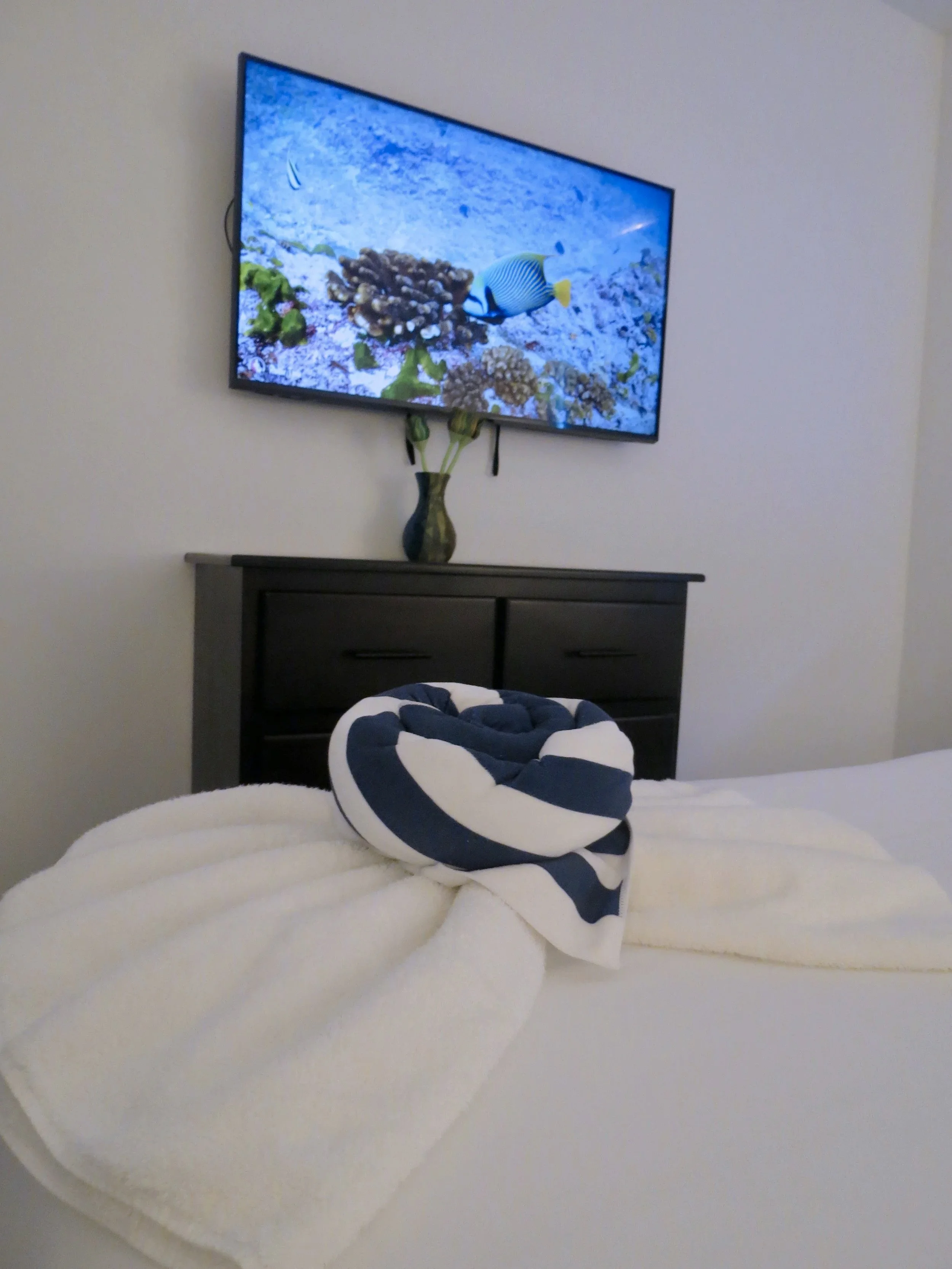 A bedroom with a television mounted on the wall displaying an underwater scene with coral and fish, a dark dresser below the TV, a vase with flowers on the dresser, and a bed with white bedding and a rolled-up navy blue and white striped towel or bla