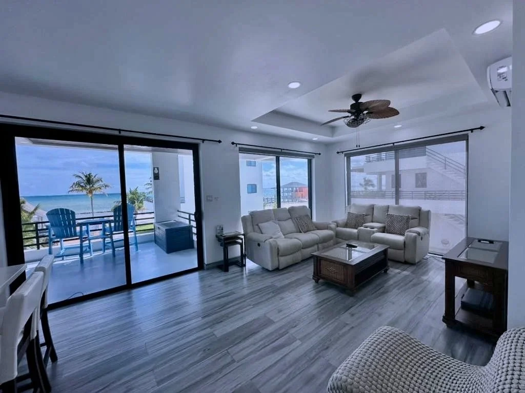 Living room with large glass sliding doors opening to a balcony with outdoor chairs and a view of the beach, ocean, and palm trees. Interior includes beige sofas, a coffee table, ceiling fan, and modern decor.  Unit S2, Caye Caulker,  Blu Zen Belize