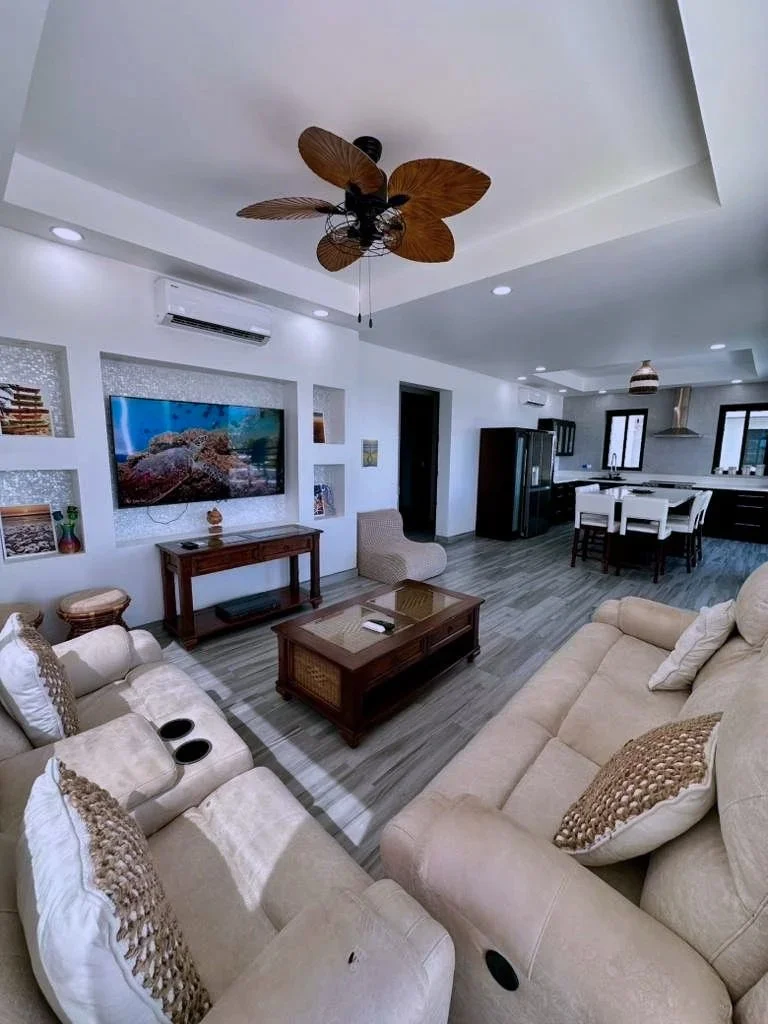 Living room with beige sofas, a wooden coffee table, wall-mounted TV, and open kitchen in the background.  Unit S2, Caye Caulker,  Blu Zen Belize
