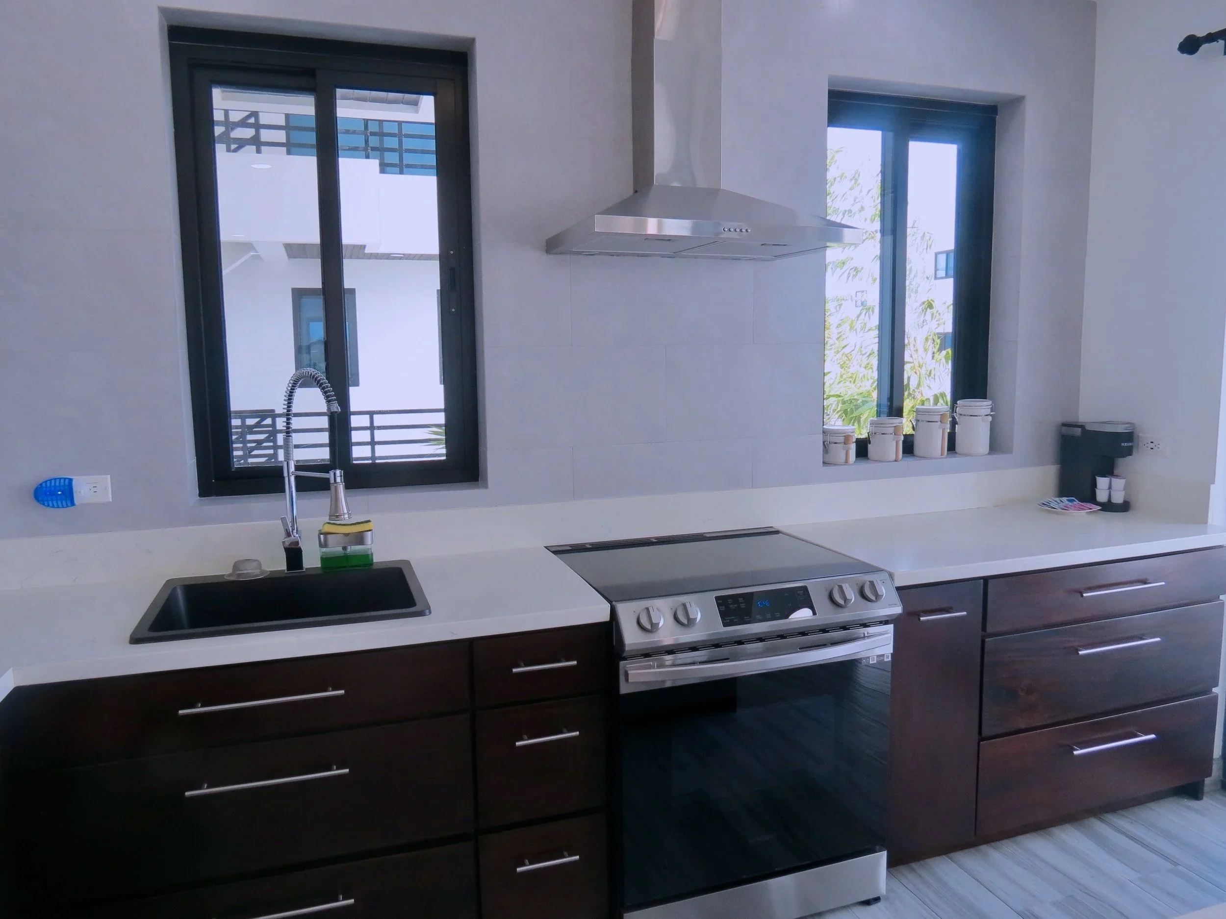 Modern kitchen with dark wood cabinets, white countertop, stainless steel stove, black sink, window with black frames, and various containers on the windowsill.  Unit S2, Caye Caulker,  Blu Zen Belize